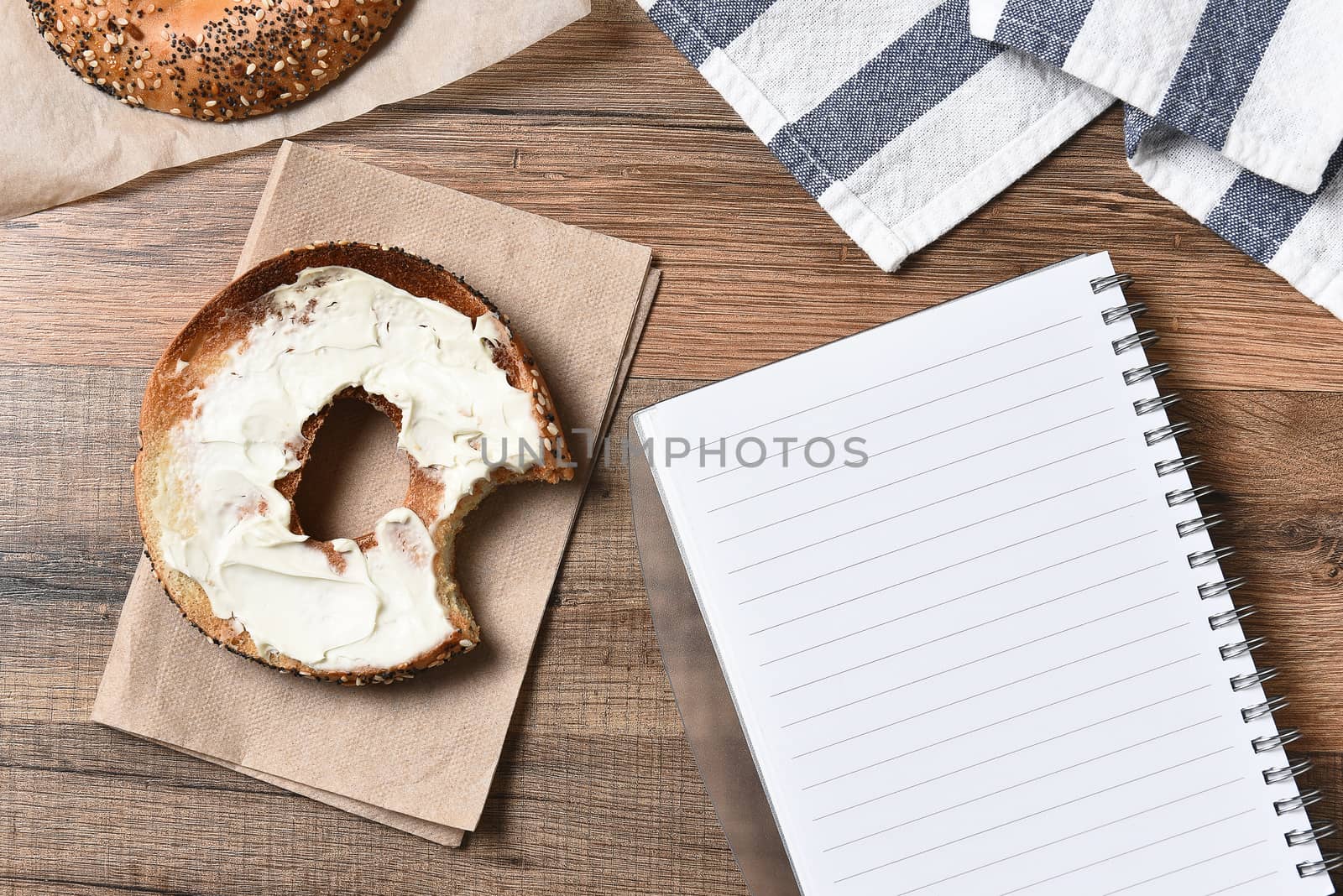 A bagel with cream cheese and a bite taken out next to a note pad. Top view in horizontal format.