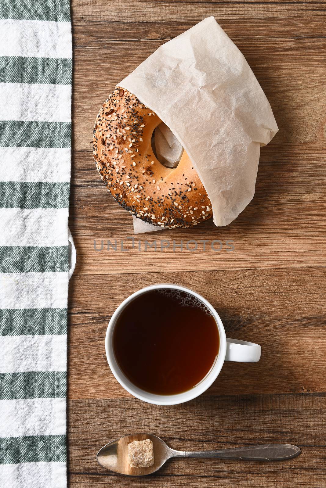 High angle view of a bagel, cup of coffee, and spoon with raw sugar lump with a striped towel.