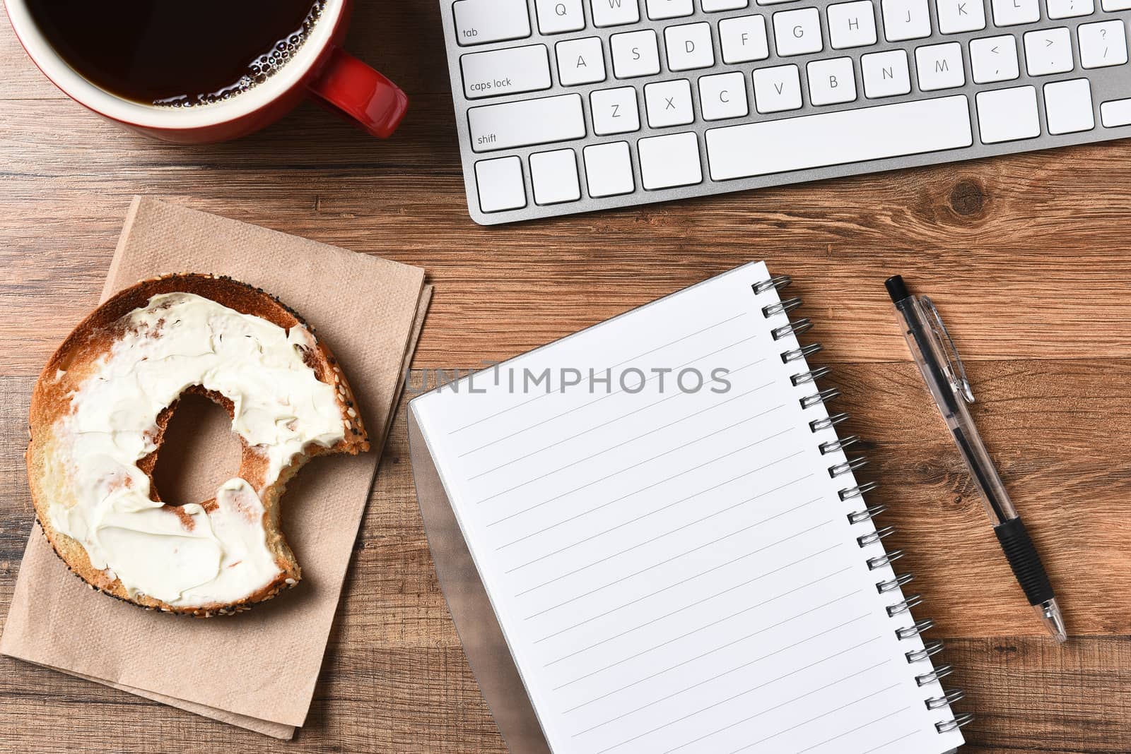 A bagel with cream cheese and a bite taken out next to a computer keyboard and a pad and pen. A cup of coffee in the upper corner, Horizontal for a high angle. 