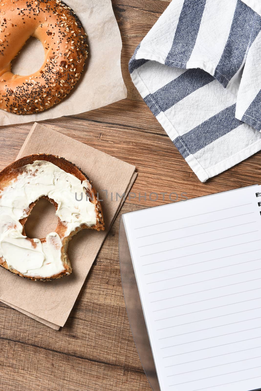 A bagel with cream cheese and a bite taken out. With a towel and note pad on a rustic wood desk. High angle view in vertical format,