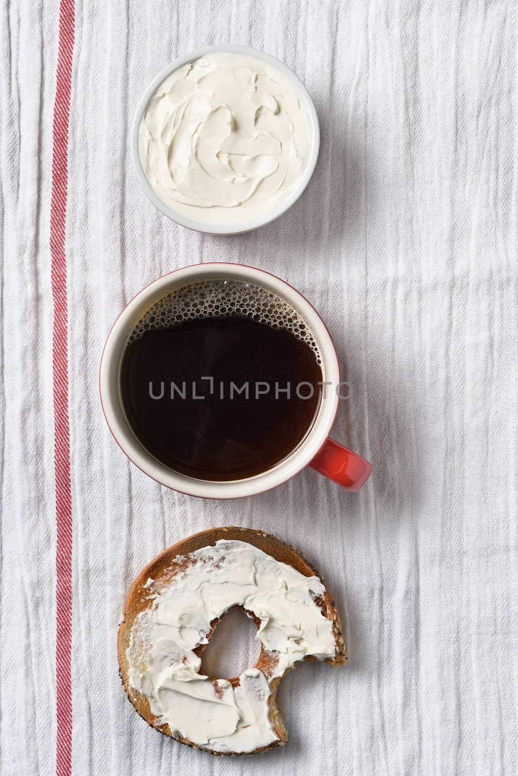 A bagel with cream cheese and a bite taken out. on a towel with cup of coffee and crock filled with cream cheese. Vertical format with copy space.