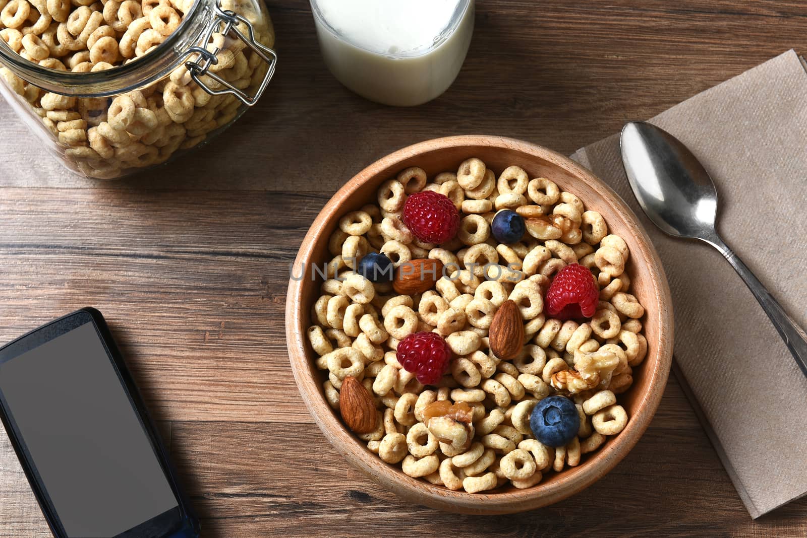 High angle view of a bowl of breakfast cereal with blueberries, raspberries and nuts. A bottle of milk and cell phone are also on the rustic wood table.