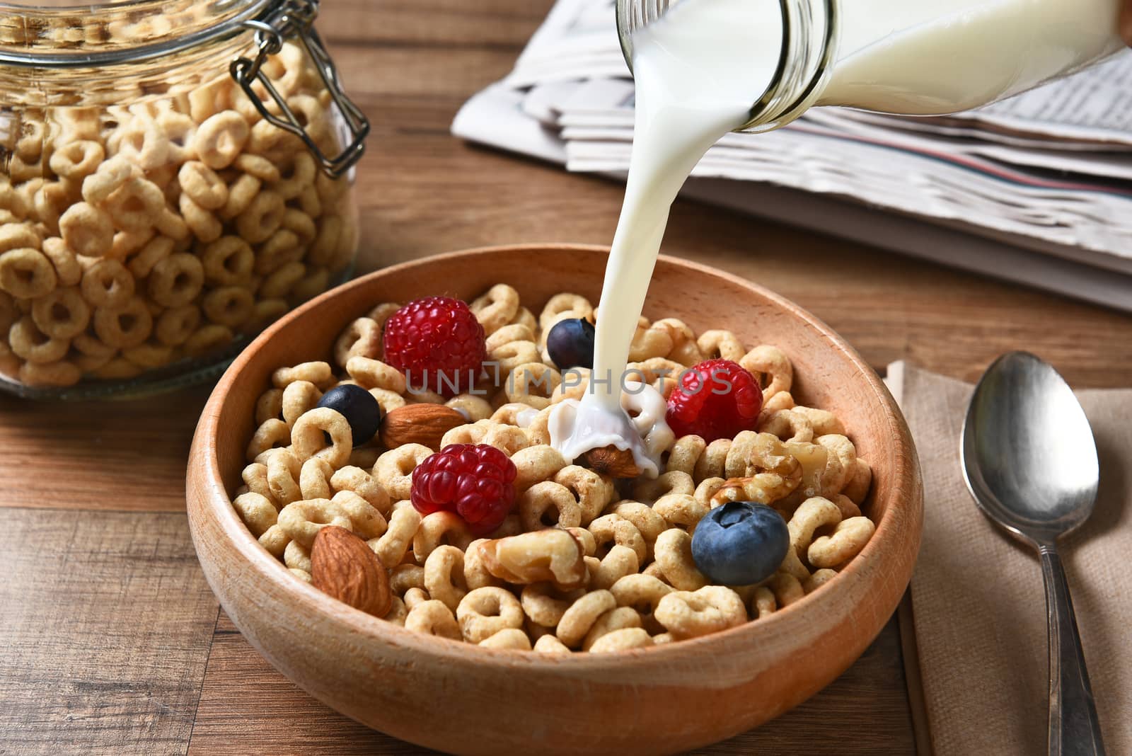 High angle view of a bowl of breakfast cereal with blueberries, raspberries and nuts. A bottle of milk is pouring into the bowl