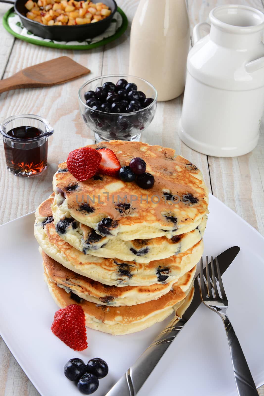 High angle shot of a stack of fresh homemade blueberry pancakes. The plate is on a rustic farmhouse style kitchen table. Other items include a syrup pitcher, knife, fork, skillet with fried potatoes, strawberries and more.
