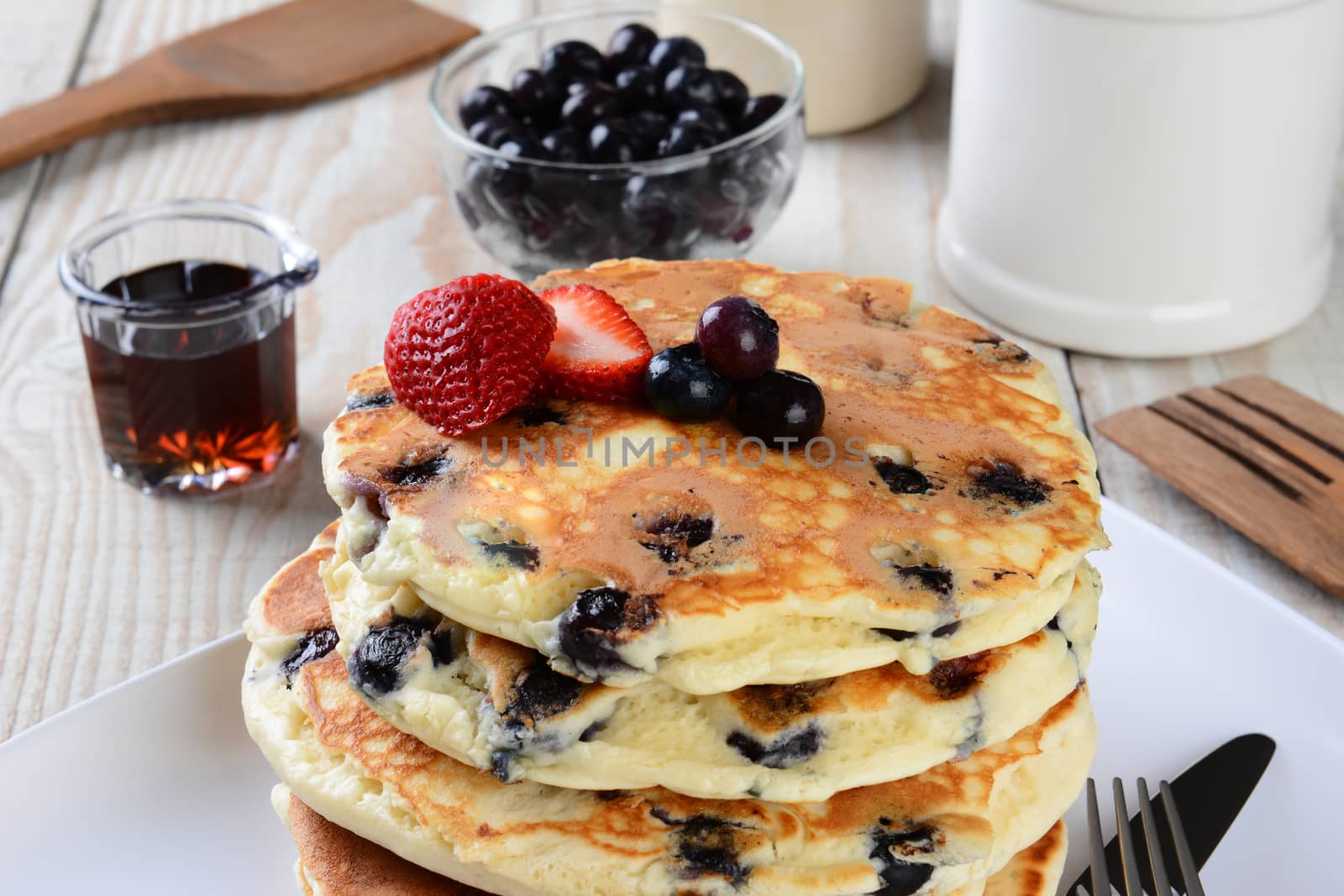 Blueberry Pancake Closeup with syrup pitcher, wooden spatula and fork, blueberry bowl and other items set out for a homemade breakfast. Horizontal format.