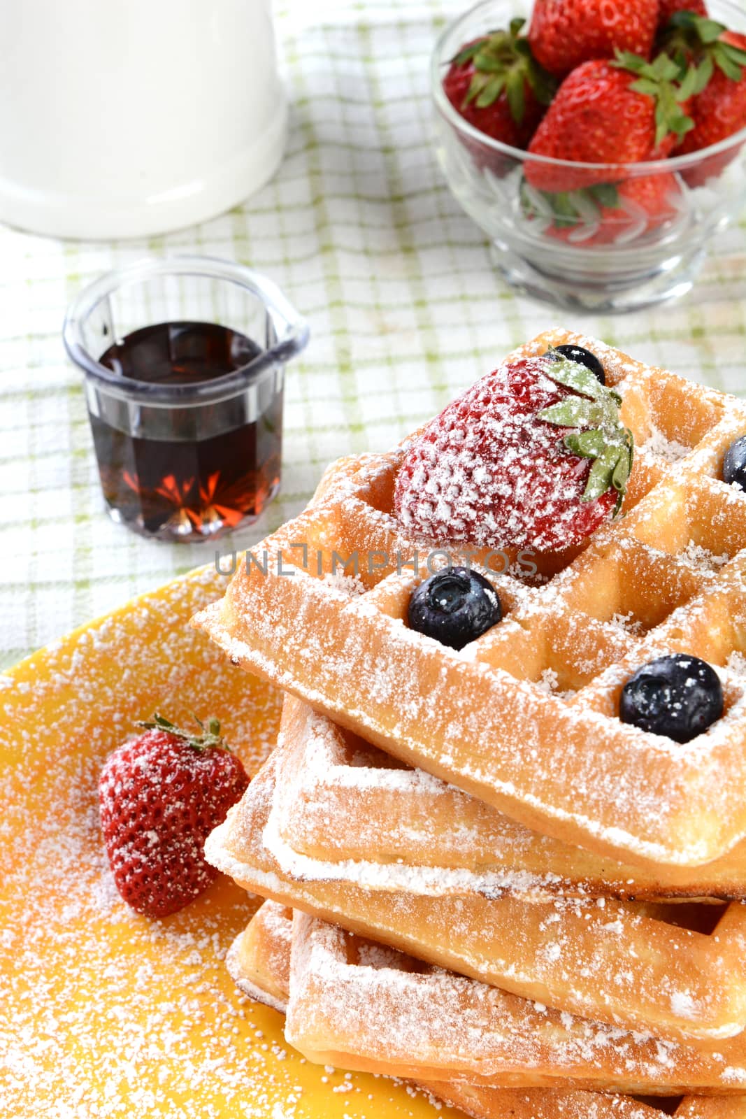 Waffles with strawberries and blueberries covered with powdered sugar. Vertical format with syrup and a bowl of fresh strawberries and a crock.