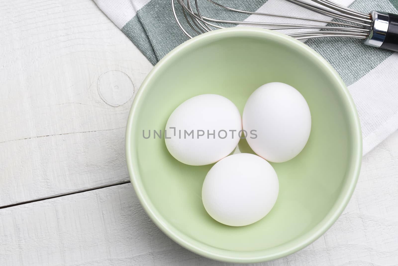 High angle view of three eggs in a bowl on a wood table with kitchen towel and whisk.