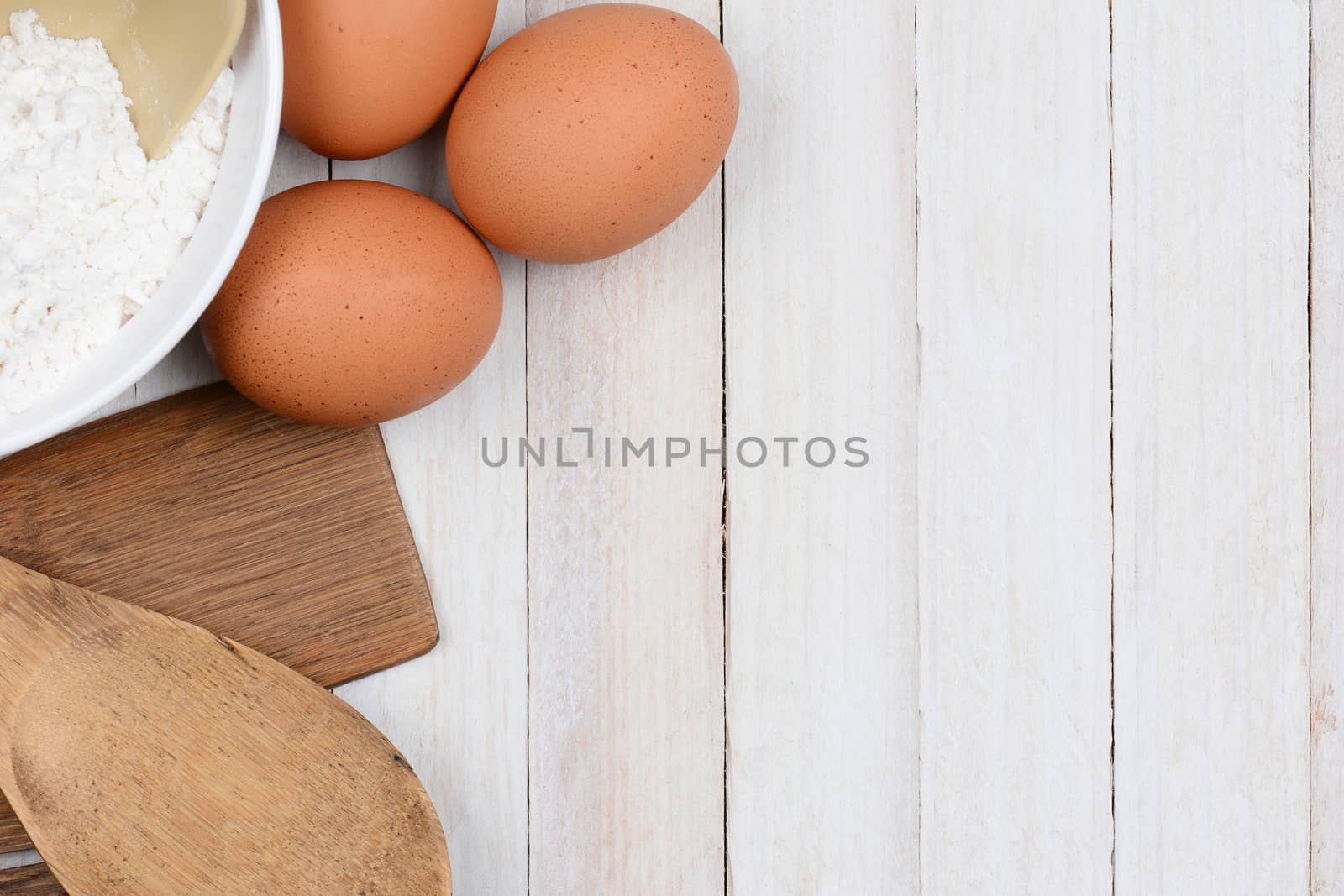 High angle image of a bowl of flour brown eggs and wood kitchen utensils on a rustic white wood table. Items are set to one side leaving copy space.