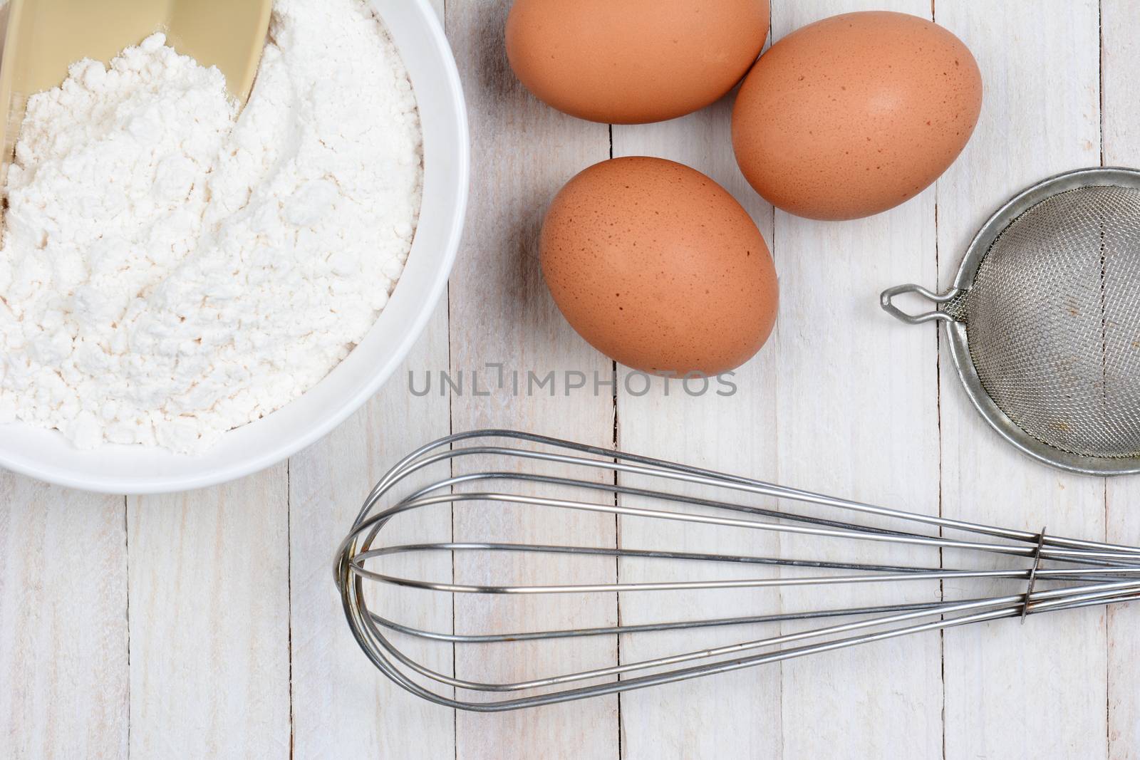 High angle closeup of three brown eggs, a bowl of flour, a whisk and strainer on a rustic white kitchen table. 