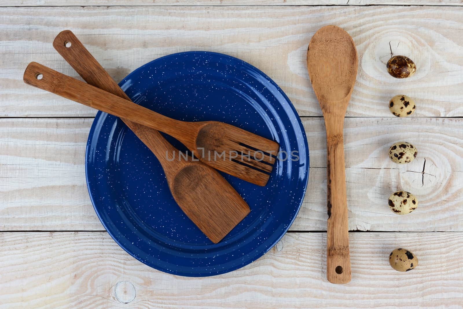 High angle shot of a blue speckled plated with wooden utensils and a line of quail eggs. Horizontal format on a rustic farmhouse style table.