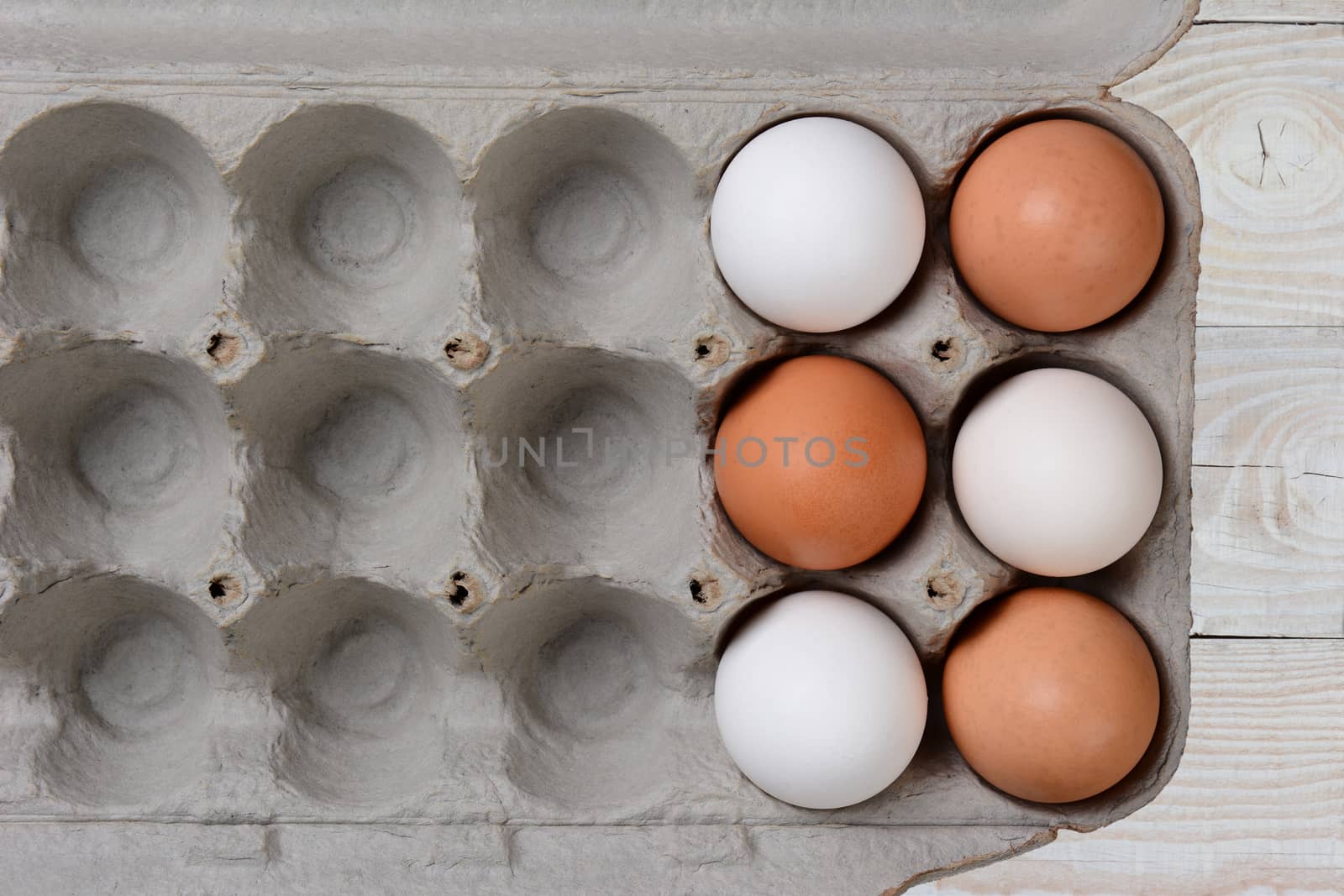 Six Eggs, three white and three brown, in a large carton with empty spaces. The carton is on a white farmhouse style wood kitchen table. 