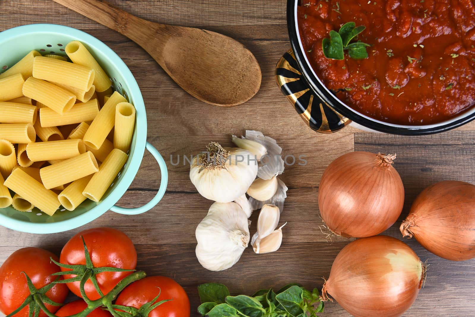 Italian meal ingredients on a wood kitchen table. A pot of sauce, colander of rigatoni, tomatoes, garlic, oregano, and onions seen from a high angle. 