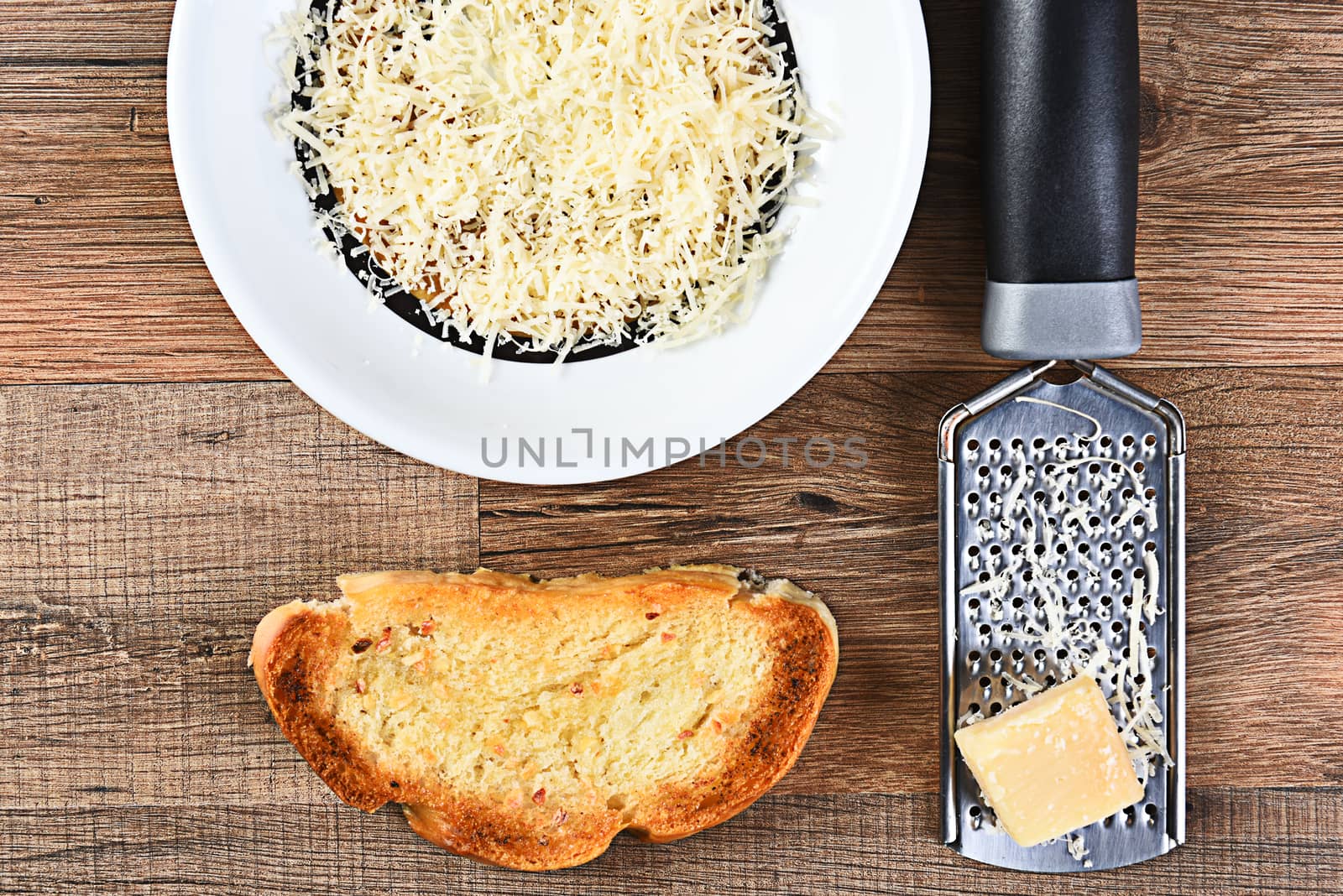 High angle view of a piece of garlic bread and a bowl of grated parmesan cheese with a grater and lump of cheese.