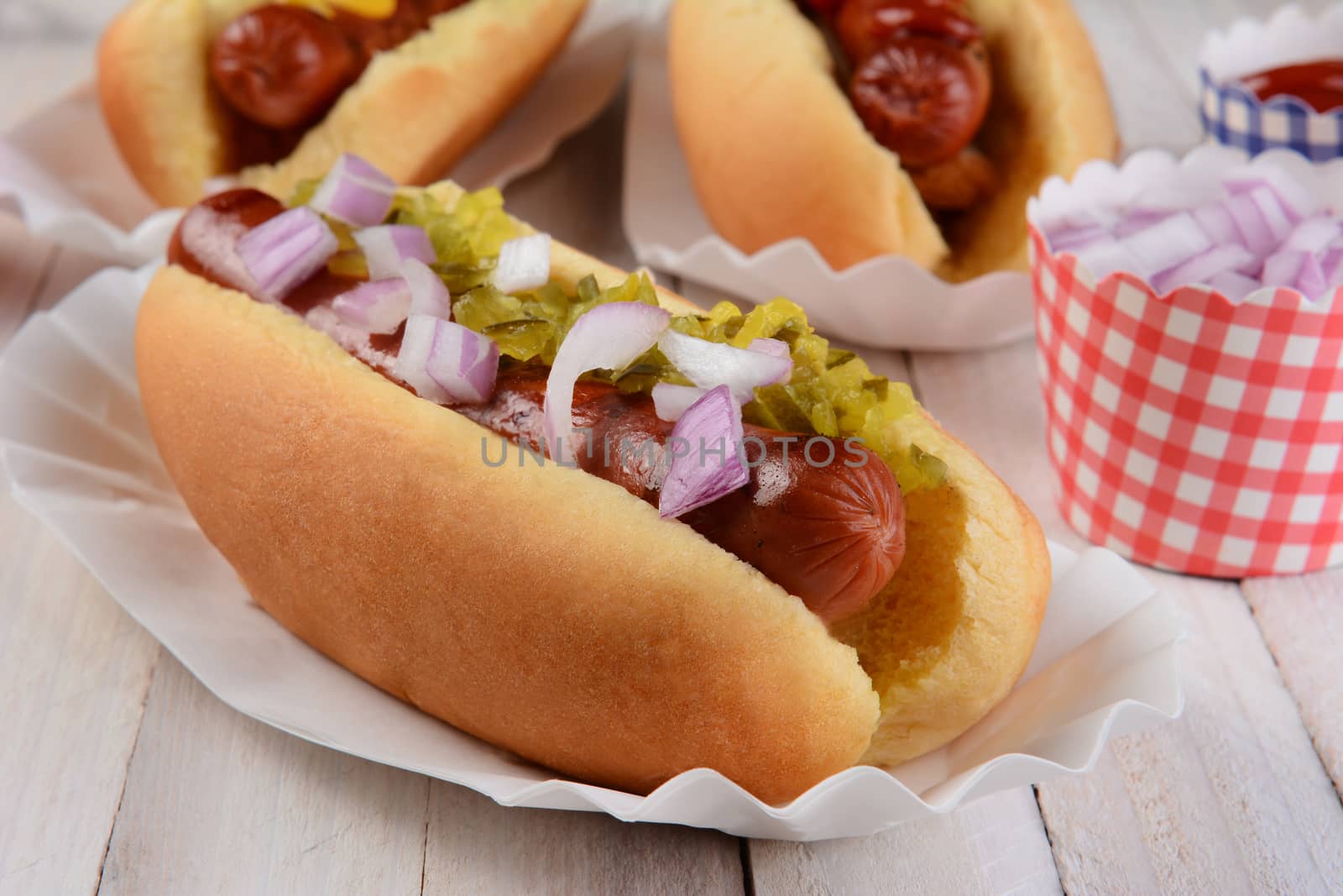 Closeup of a hot dog with relish and onions, in the background are two additional franks in buns and cups of condiments.