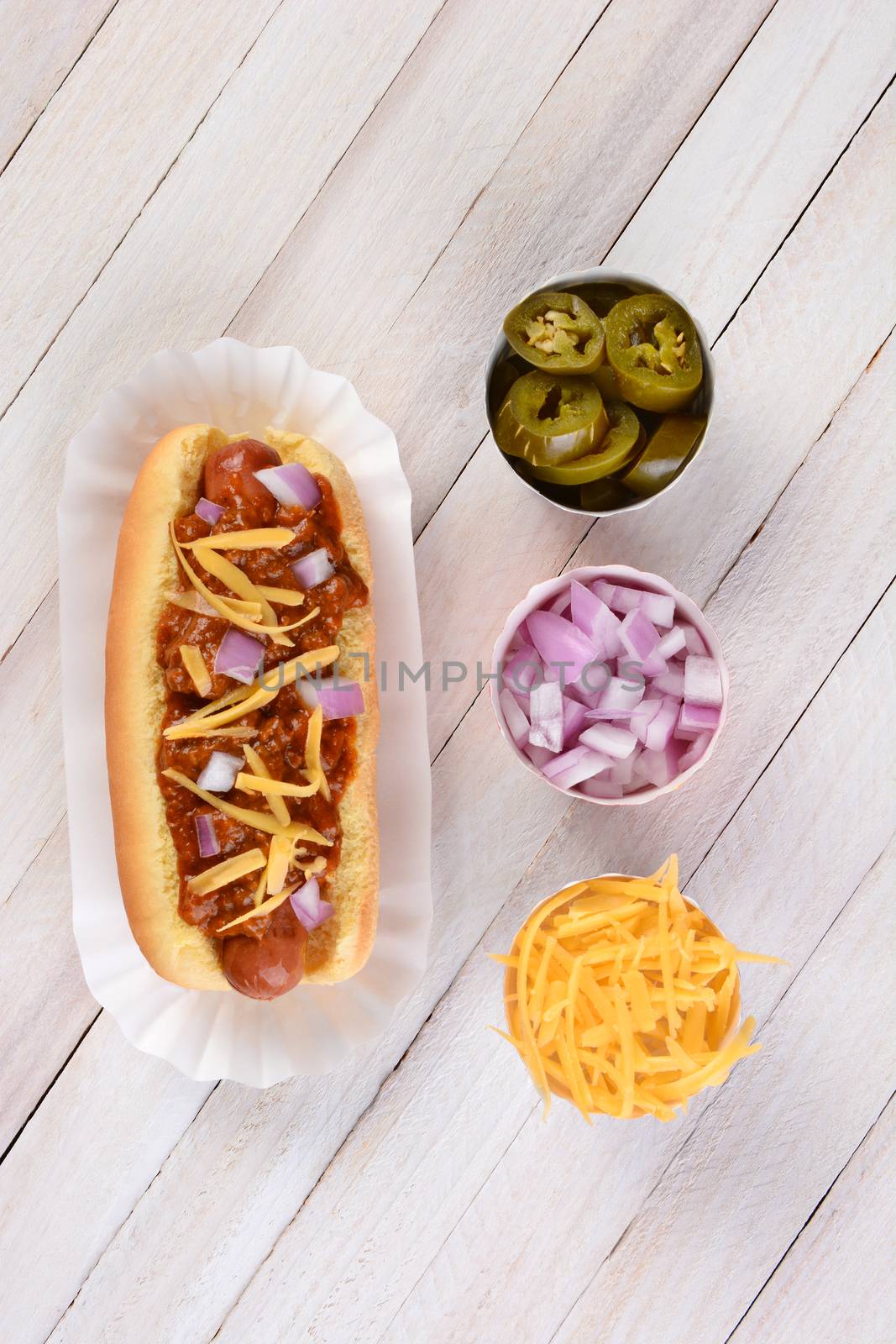 High angle shot of a Chili Hot Dog with chili, onions and shredded cheese on a rustic white wood picnic table. Cups of chopped onions, peppers and cheese are lined up by its side.