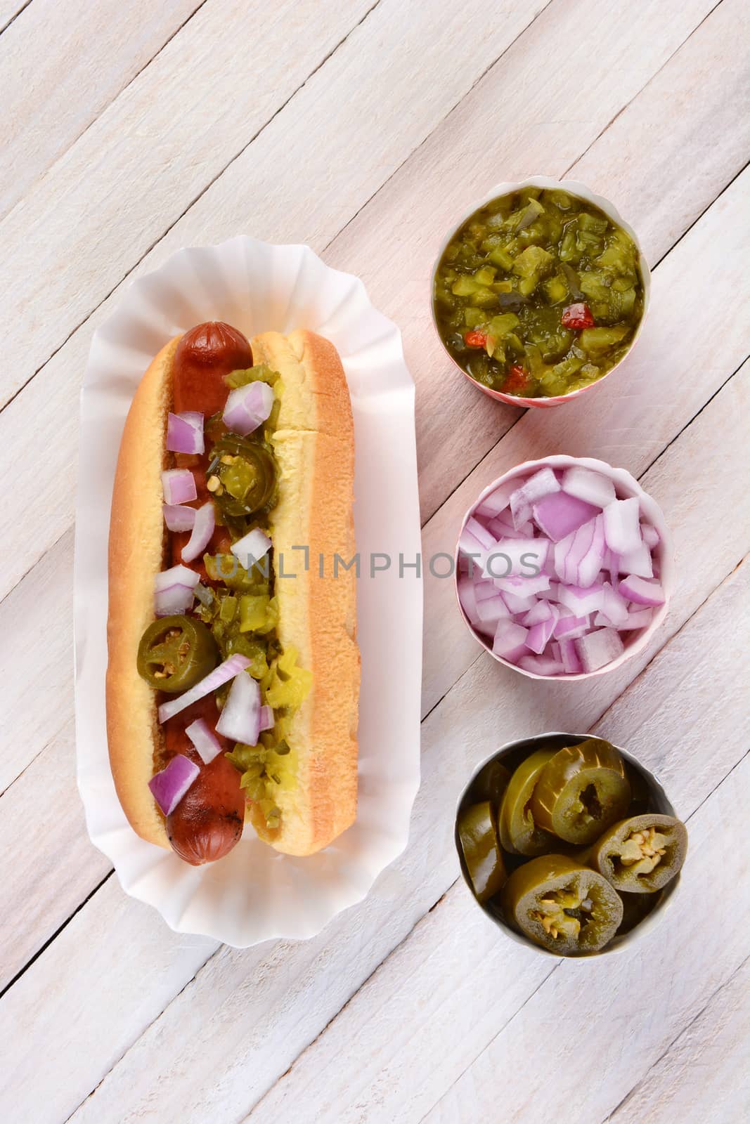 Overhead still life of a summer picnic table with a hot dog smothered in relish onions and jalapenos. Three cups of condiments are lined up next to the sandwich.