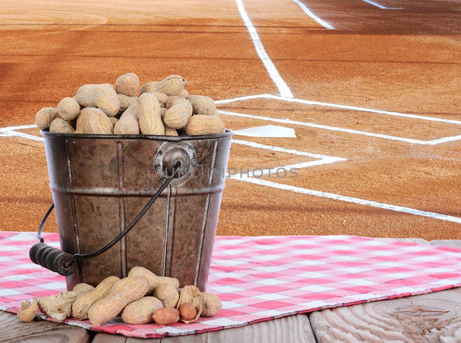 Closeup of a pail full of peanuts on a wood picnic table and a red checked tablecloth. Horizontal format with a baseball field background with copy space