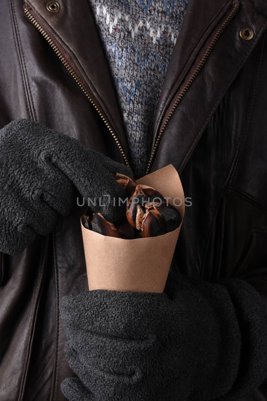 Closeup of a man in sweater and leather jacket holding a brown paper cone of fresh roasted chestnuts.