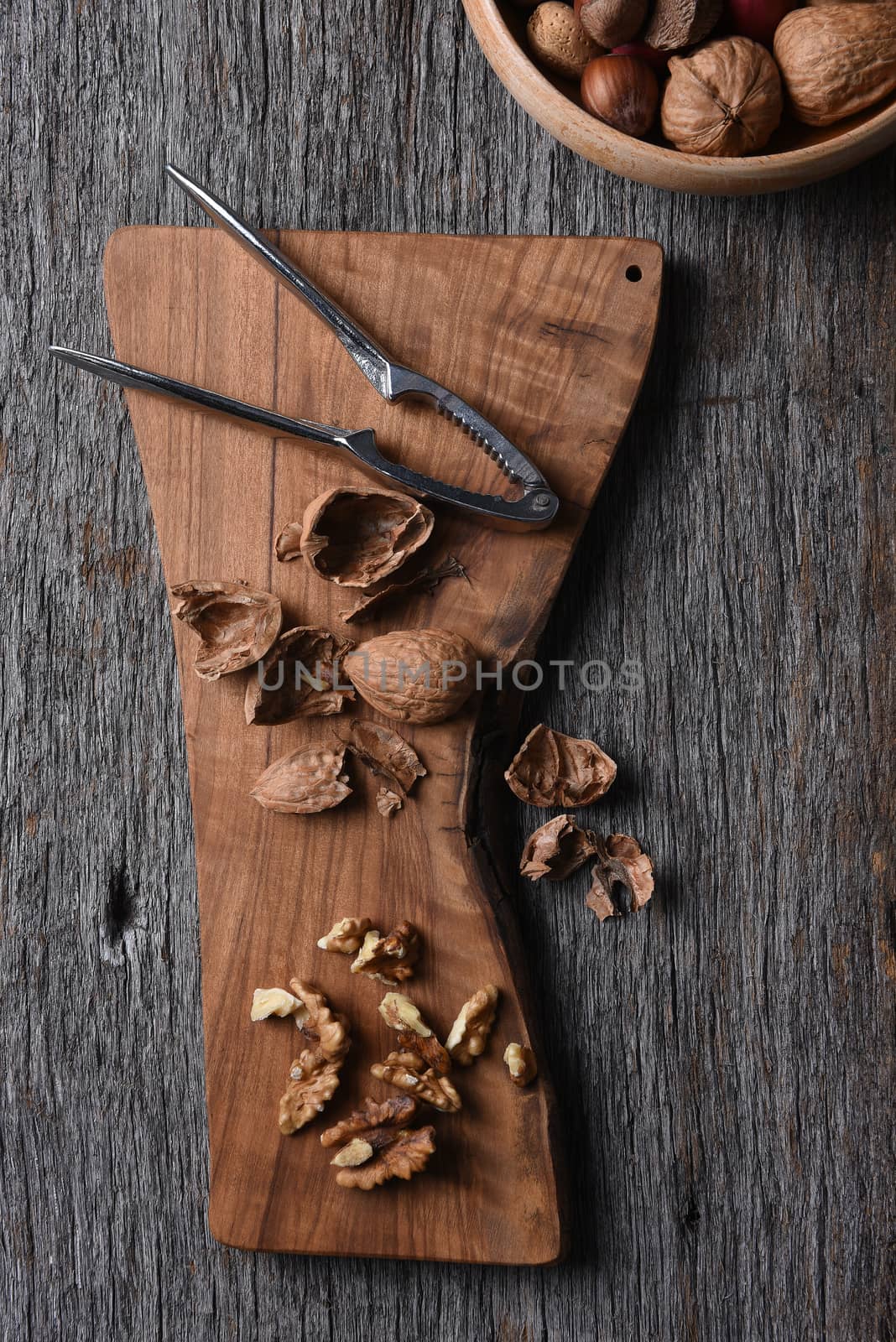 Cutting board with cracked walnuts and nutcracker with a bowl of mixed nuts int he corner.