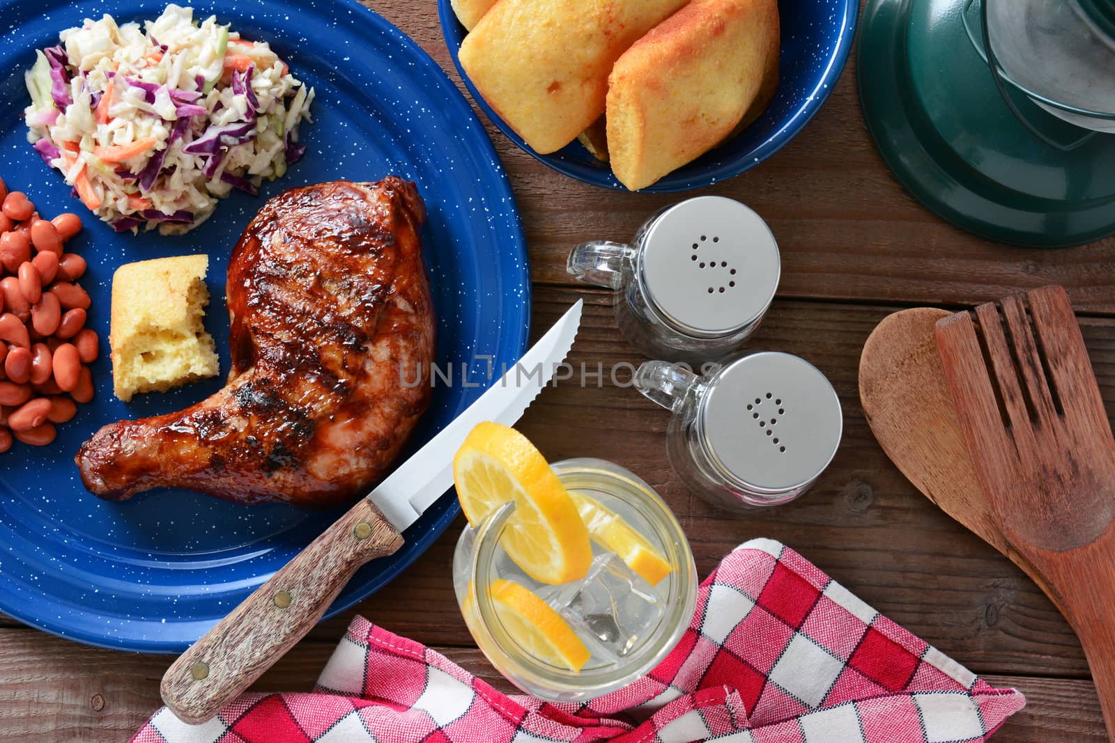 High angle shot of a barbecue chicken plate with cole slaw, pinto beans and corn bread. The meal is on a rustic wooden restaurant table with a red and white checked napkin and a glass of lemonade.