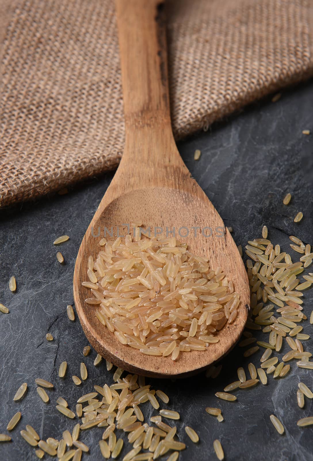 Closeup of a wood spoon with brown rice, on a slate background and burlap sack. 