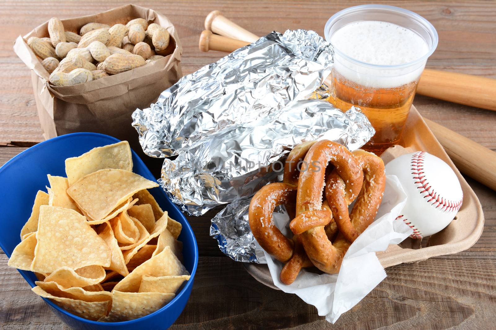 Closeup of a tray of food and souvenirs that one would find at a baseball game. Items include, hot dogs wrapped in foil, beer, peanuts, chips, baseball, mini bats and pretzels. Horizontal format.