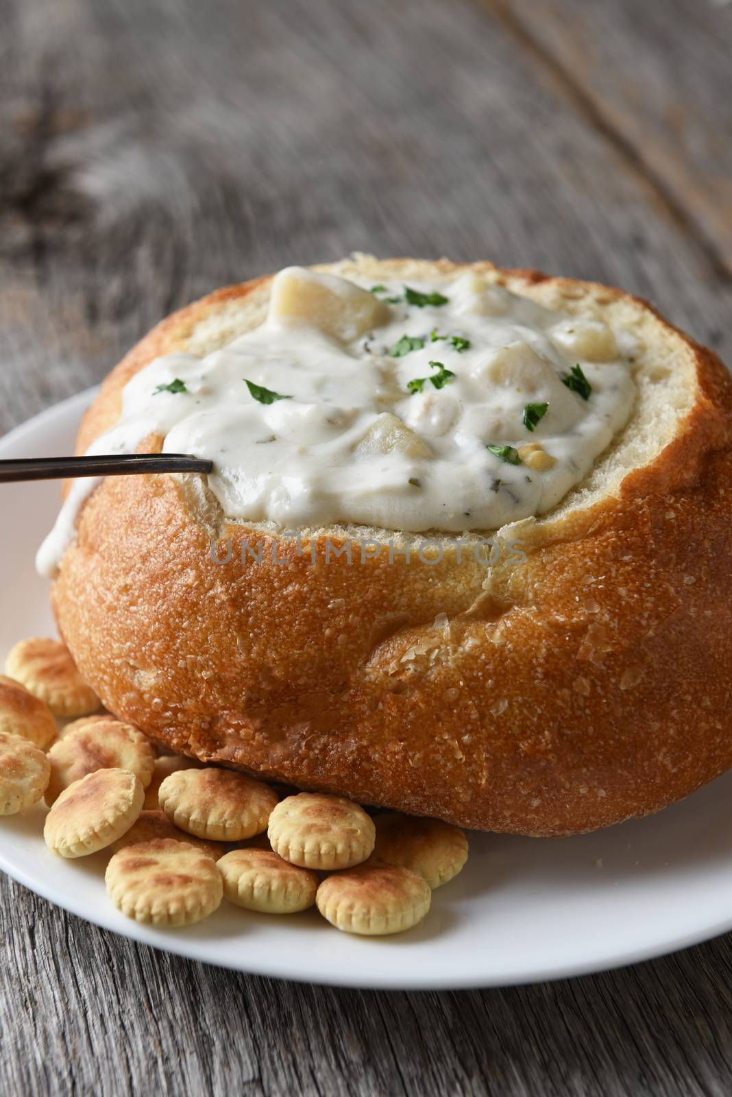 Vertical closeup of a bread bowl of New England Clam Chowder on a rustic wood table with a spoon and oyster crackers.