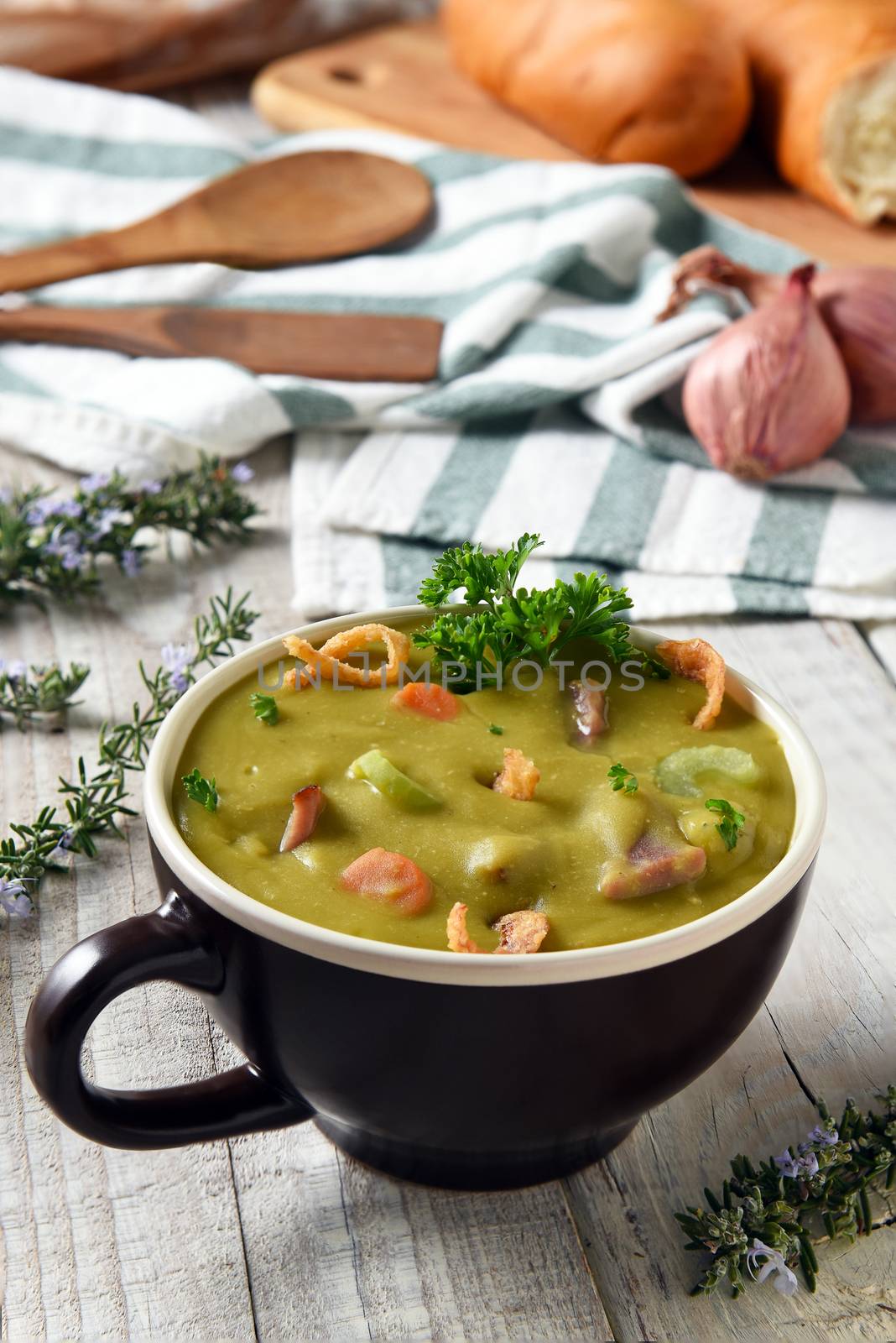 Split Pea Soup with ham and vegetables in a mug on a white rustic kitchen table with bread, towel, wood utensils and herbs.