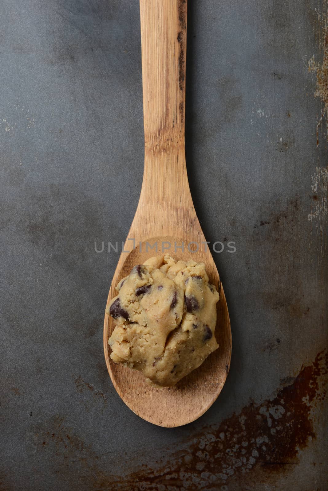 High angle closeup of a gob of chocolate chip cookie dough on a wooden spoon in the middle of an old used cookie baking sheet. Vertical format.