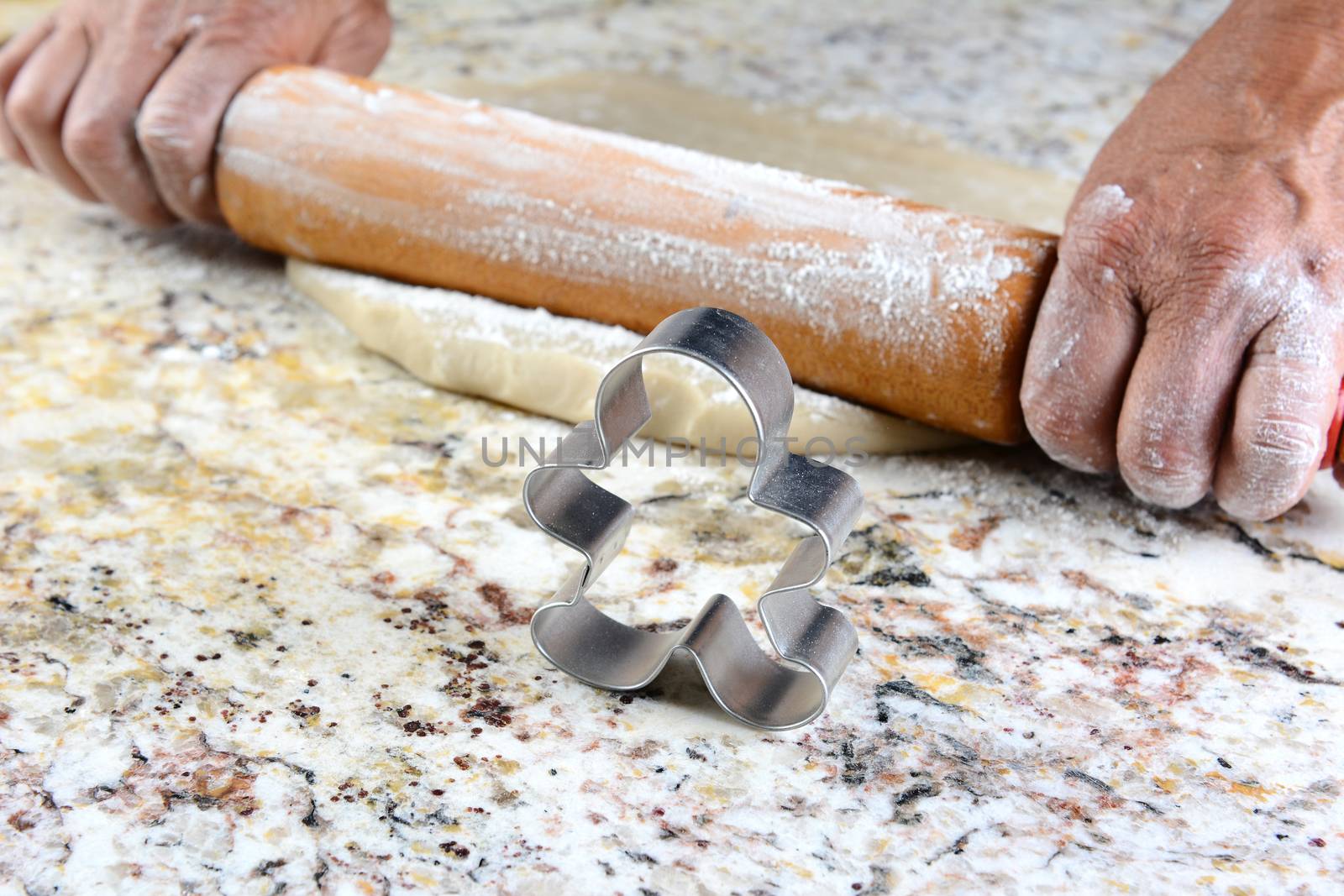 Closeup of a Gingerbread man cookie cutter on a granite counter. In the background are the hands of a baker rolling out dough. Shallow depth of field with the focus on the cookie cutter.