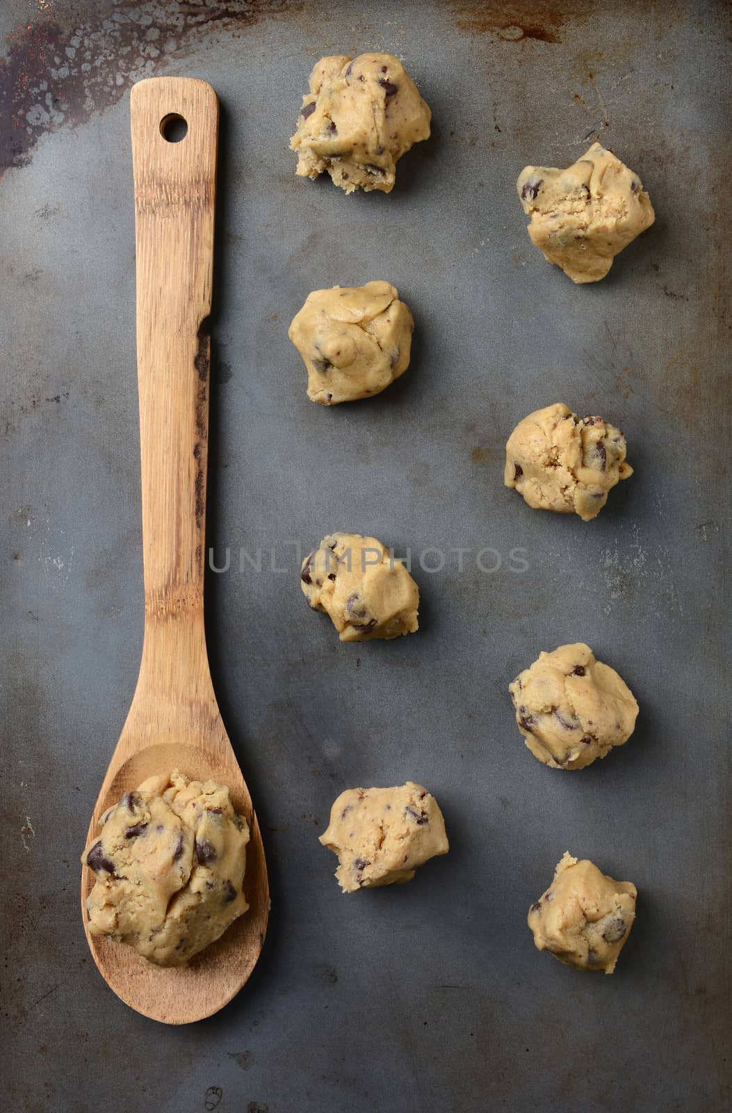 Overhead shot of chocolate chip cookie dough on a cookie sheet, with a wooden spoon holding a big gob of the raw dough. Vertical Format.