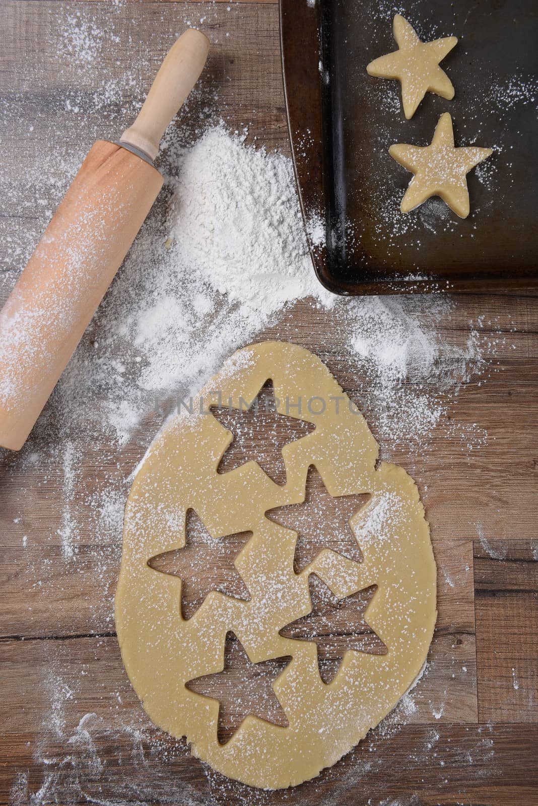 Top view of raw cookie dough with star shapes and cookie cutter on wood table with a rolling pin and flour sprinkles and baking sheet.