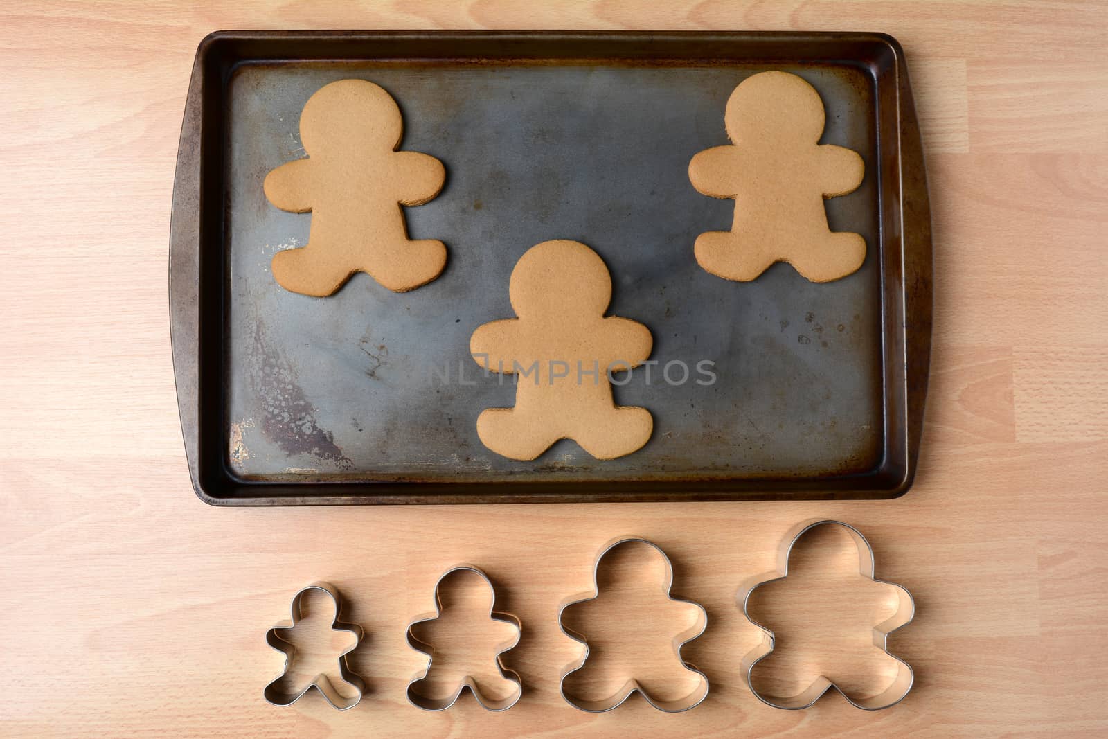 High angle shot of a cookie sheet with three gingerbread man cookies. Below the pan is a row of four cookie cutters of different sizes, Horizontal format on a butcher block kitchen table.