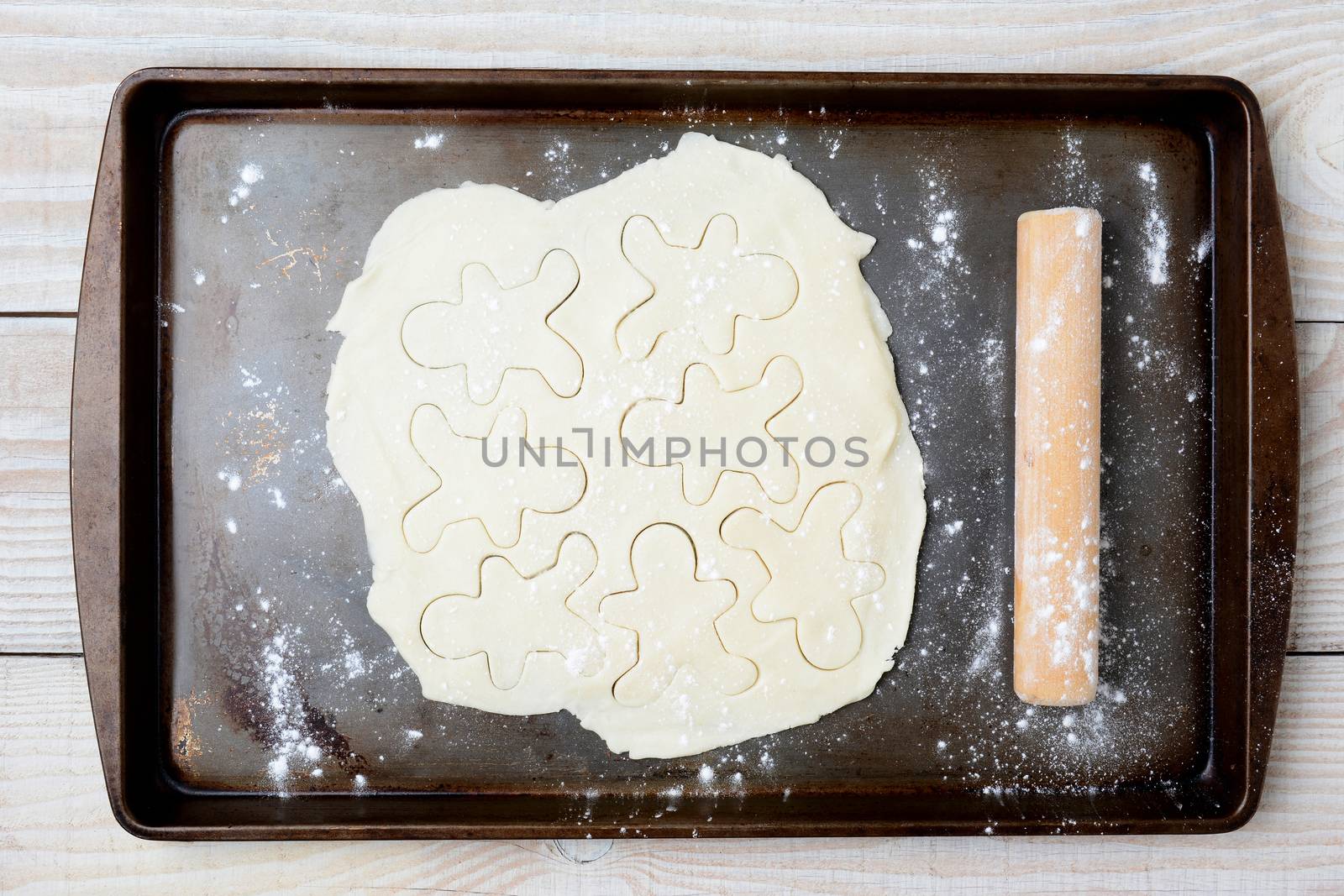 High angle shot of cookie dough with gingerbread men shapes. The dough is on an old used baking sheet with flour and a wood rolling pin. Horizontal format.