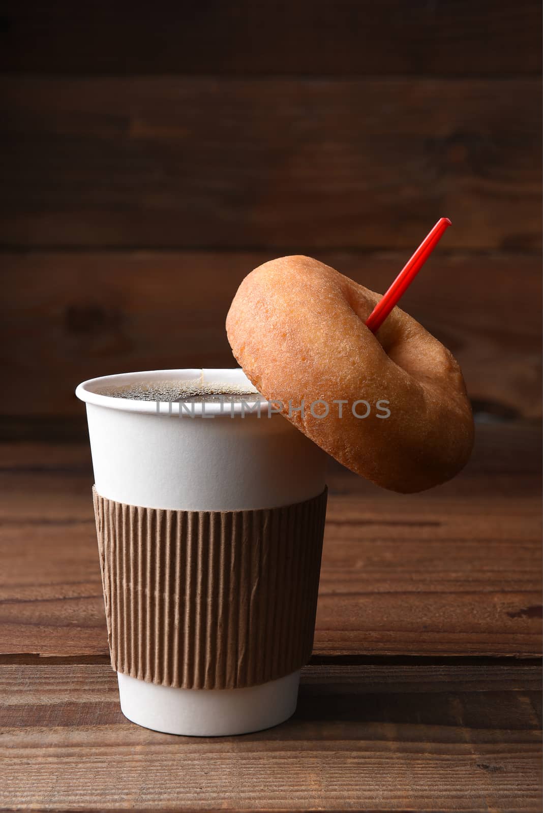 Closeup of a paper cup of hot fresh brewed coffee with a red spoon, A plain old fashioned donut is hanging on the spoon. Vertical format with copy space.