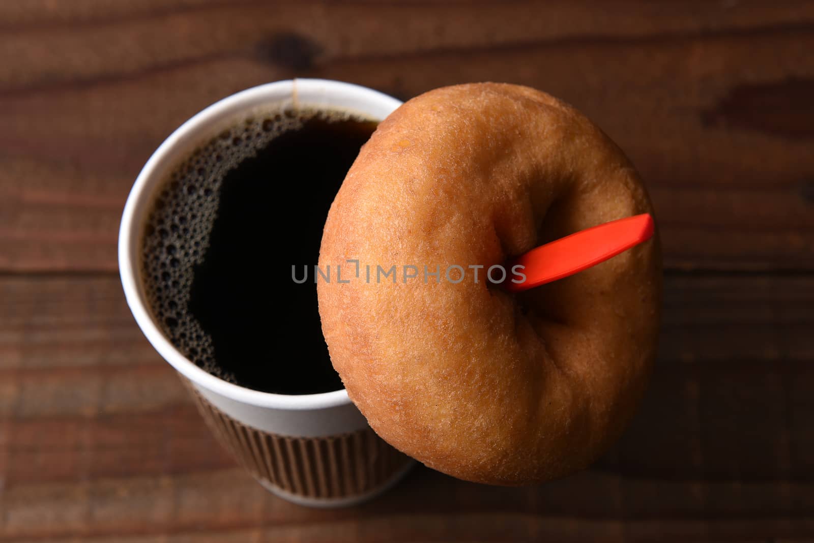 High angle view of a paper cup of hot fresh brewed coffee with a red spoon. A plain old fashioned donut is hanging on the spoon.