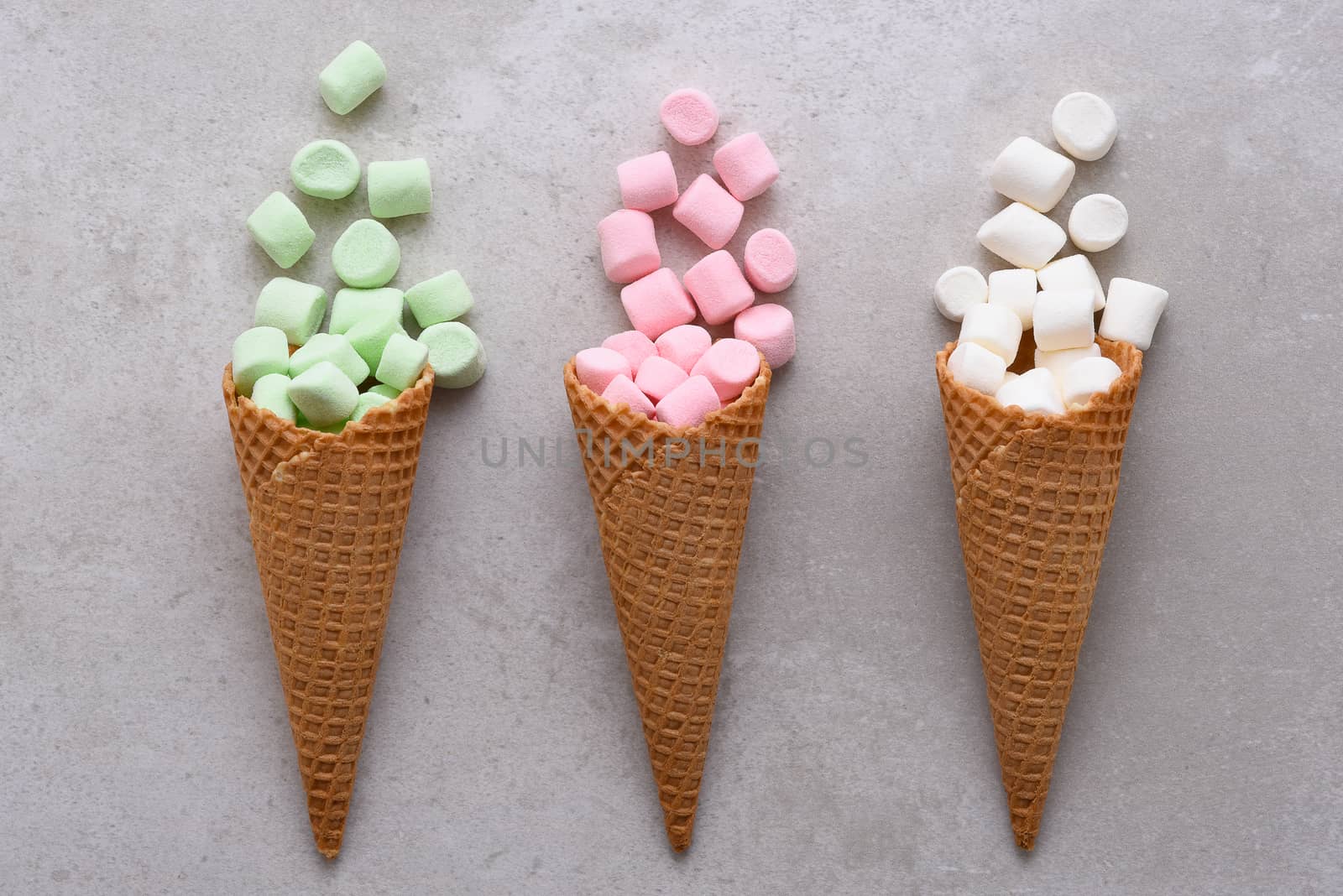 Closeup of three Sugar Waffle Ice Cream Cones filled with colorful marshmallows spilling onto tile table. 