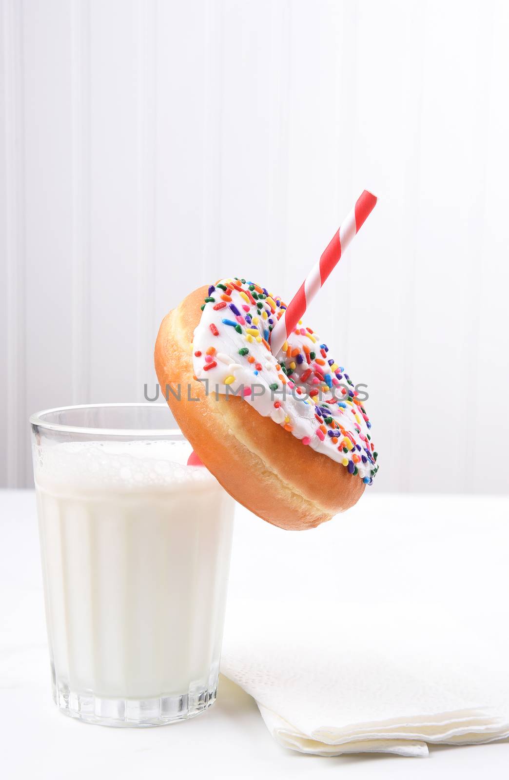 Closeup of a glass of milk with drinking straw going through the hole of a sprinkle covered donut. 