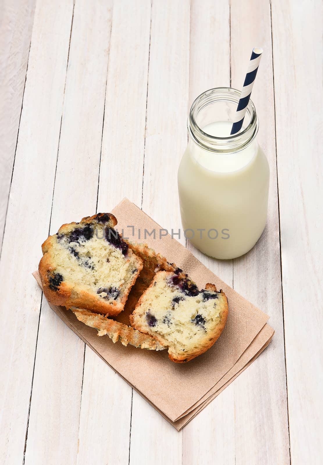 High angle view of a blueberry muffin and bottle of milk on a rustic white table. The muffin is broken in half on a napkin. Vertical format.