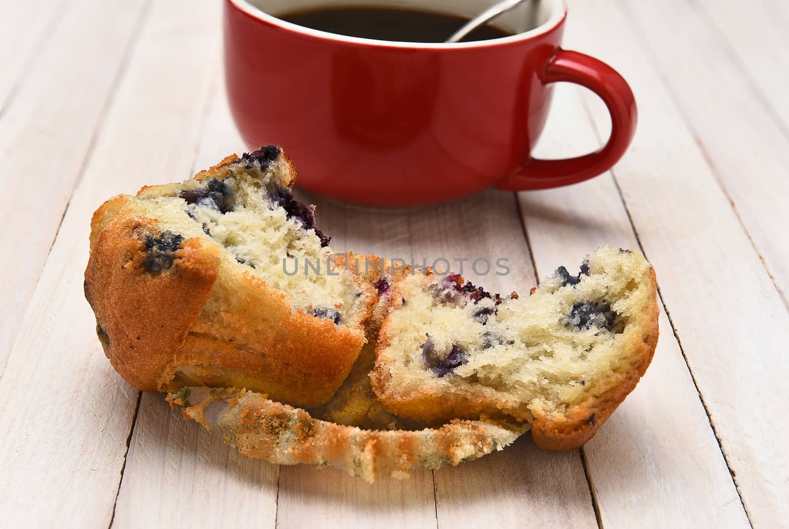 Closeup of a blueberry muffin with a cup of coffee in the background. The muffin is broken in half on a rustic white wood table.