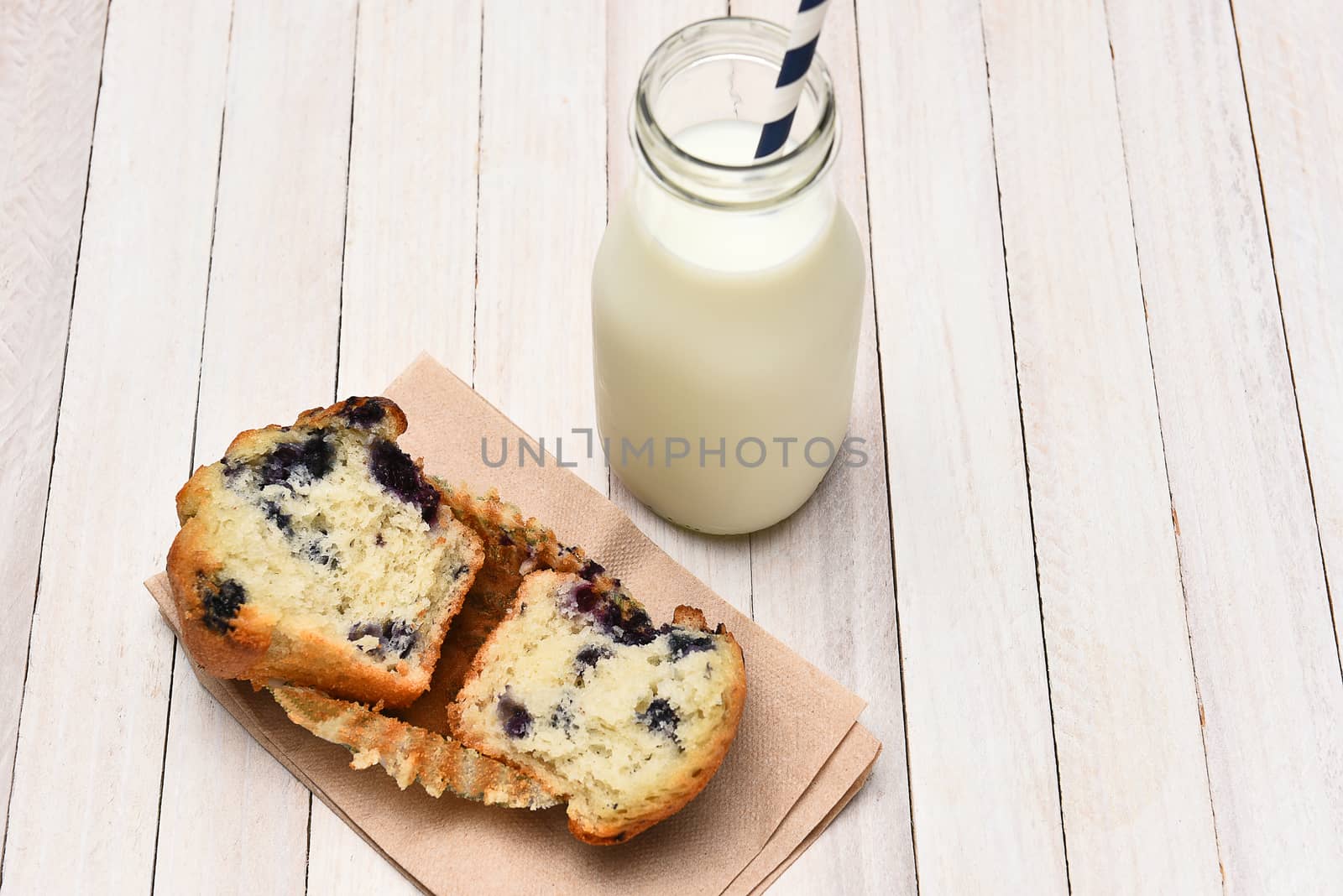 High angle view of a blueberry muffin and bottle of milk on a rustic white table. The muffin is broken in half on a napkin.