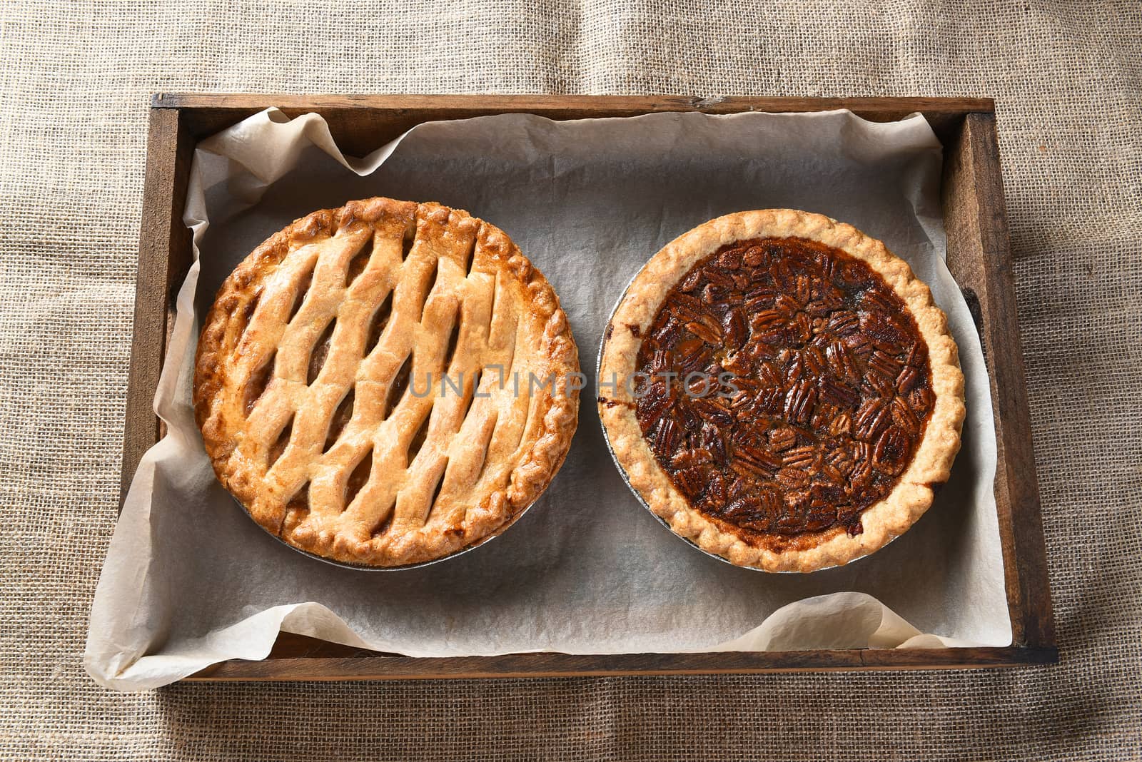 High angle view of an apple pie and pecan pie in a wood box lined with parchment paper. Horizontal format on a burlap table cloth.