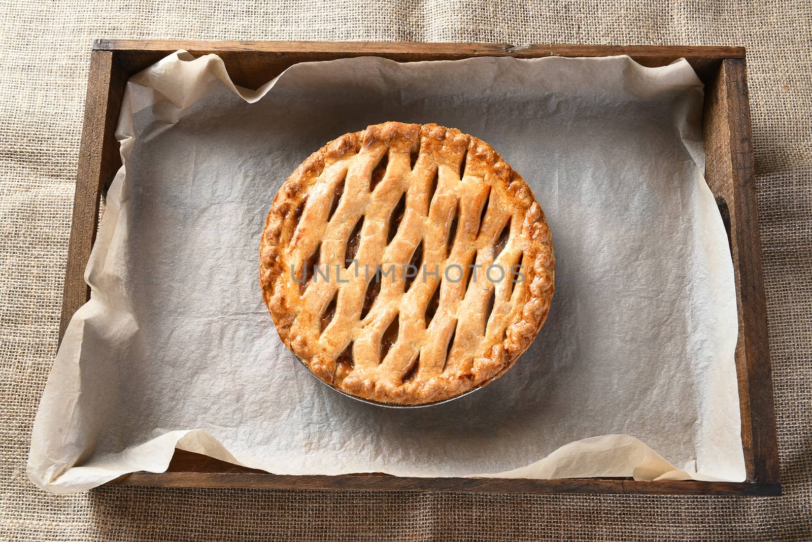 Overhead view of an Apple pie in a wood box with parchment paper. The fresh baked pie is cooling on a table with a burlap table cloth.
