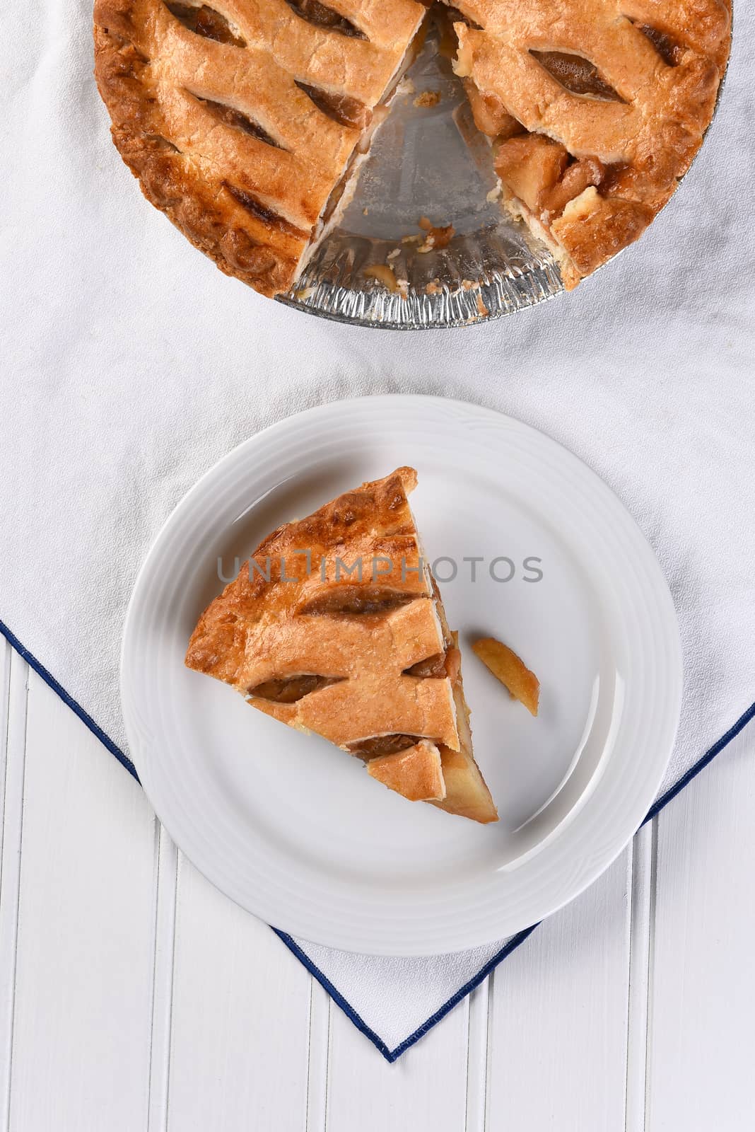 Overhead view of a slice of apple pie on a plate the rest of the pie is at the top of the frame. Vertical fomat on a white towel.