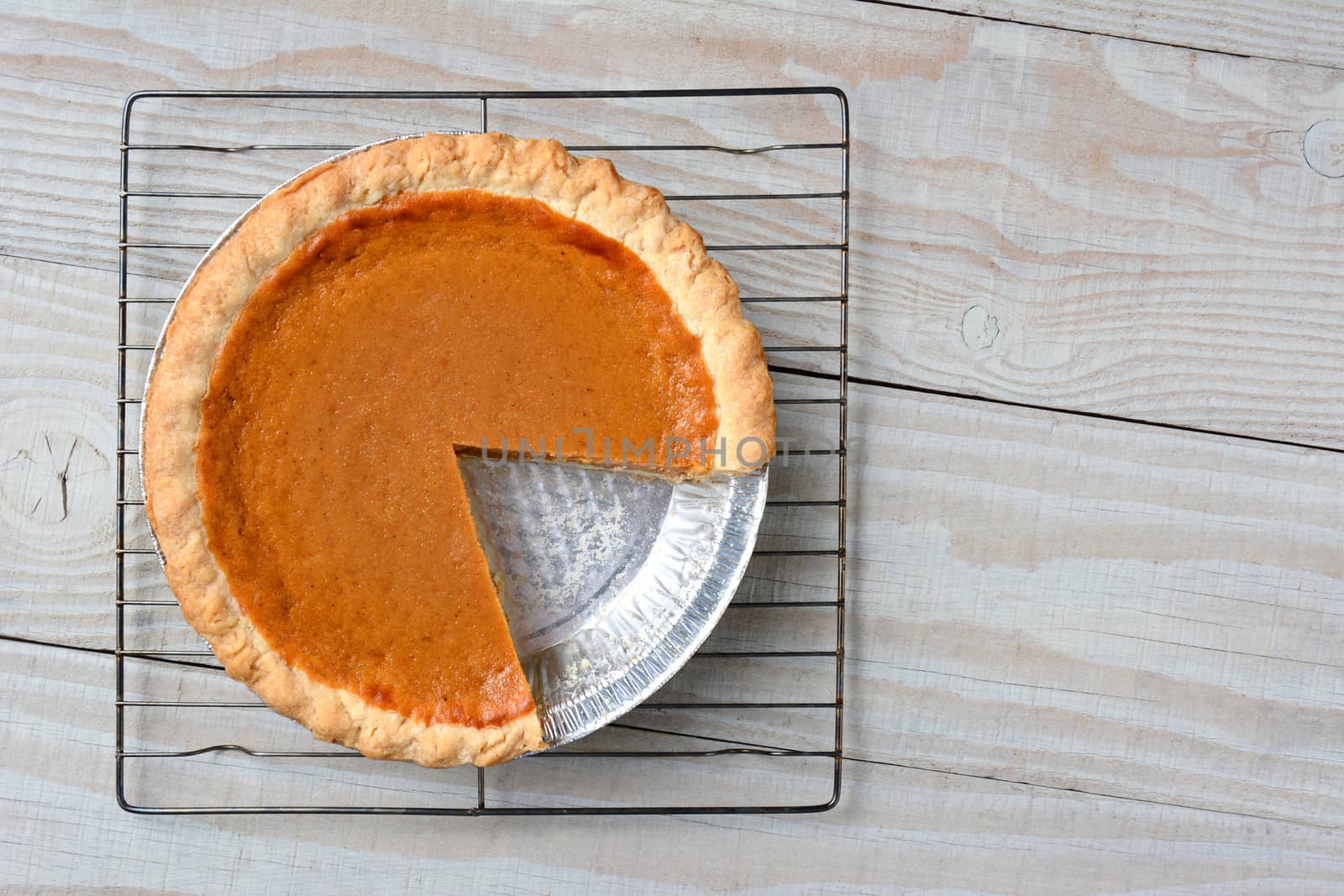 Overhead shot of a pumpkin pie with a slice cut out on a cooling rack. Horizontal format on a rustic whtie kitchen table.