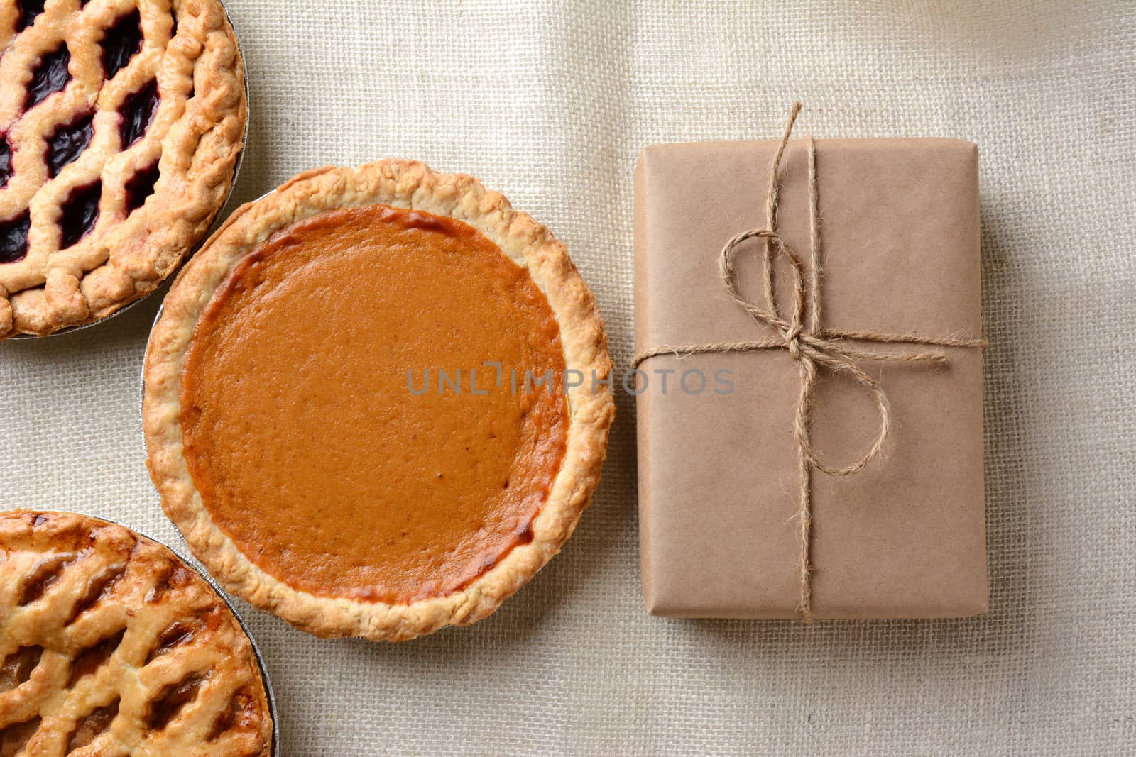 High angle shot of three holiday pies and a wrapped parcel on a burlap tablecloth. The cherry and apple pies run out of the frame with a whole pumpkin pie and package dominate the frame.