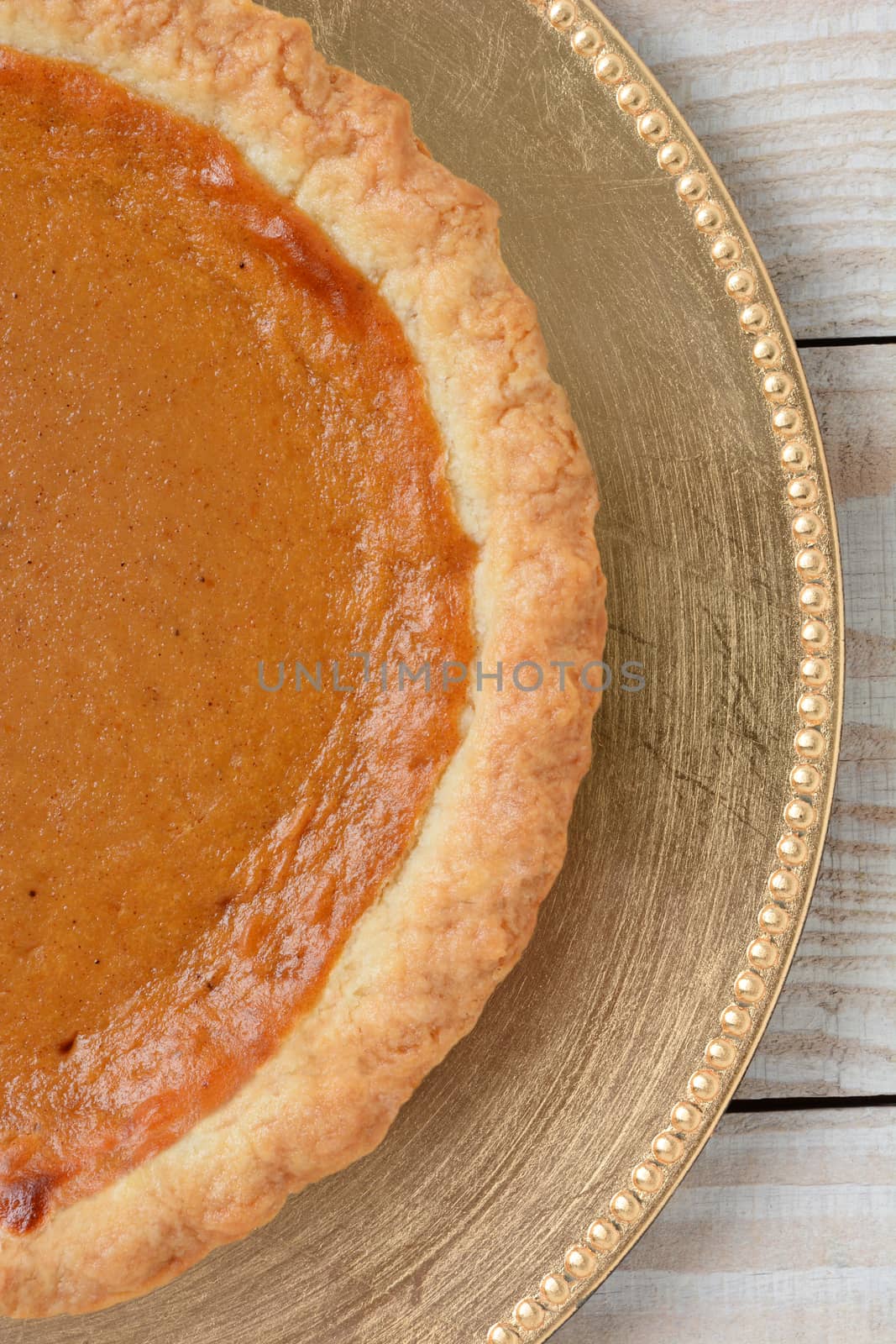 High angle closeup of an fresh baked pumpkin pie on a holiday plate. The dessert is on a gold charger on a white wood table. Vertical format.