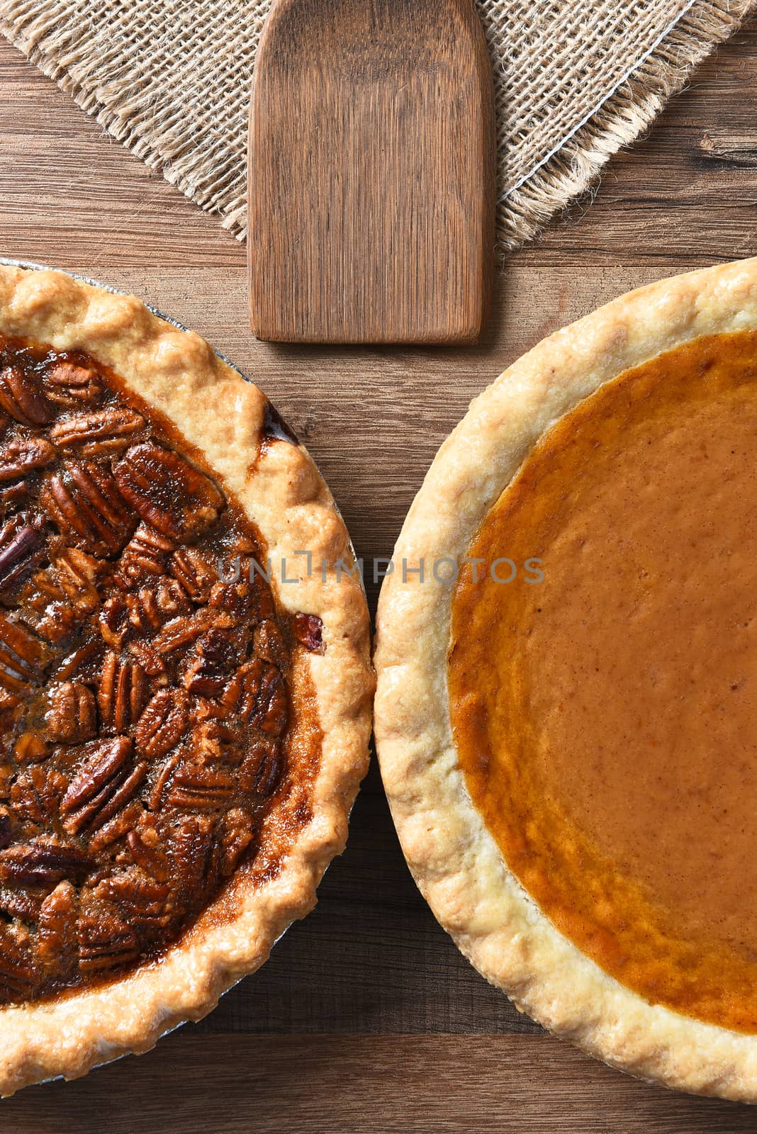 High angle vertical of half of a Pecan Pie and a Pumpkin Pie. The fresh baked pies are on a wood table with burlap table cloth and wood utensil.