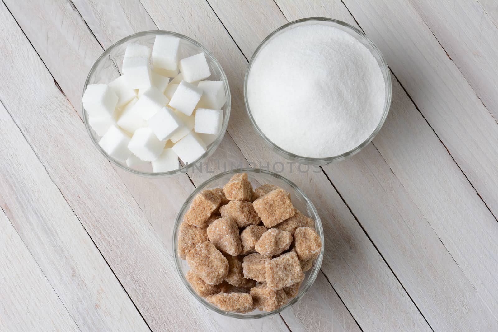 Three glass bowls filled with granulated sugar, white sugar cubes and natural sugar chunks. High angle view on a rustic wood kitchen table.