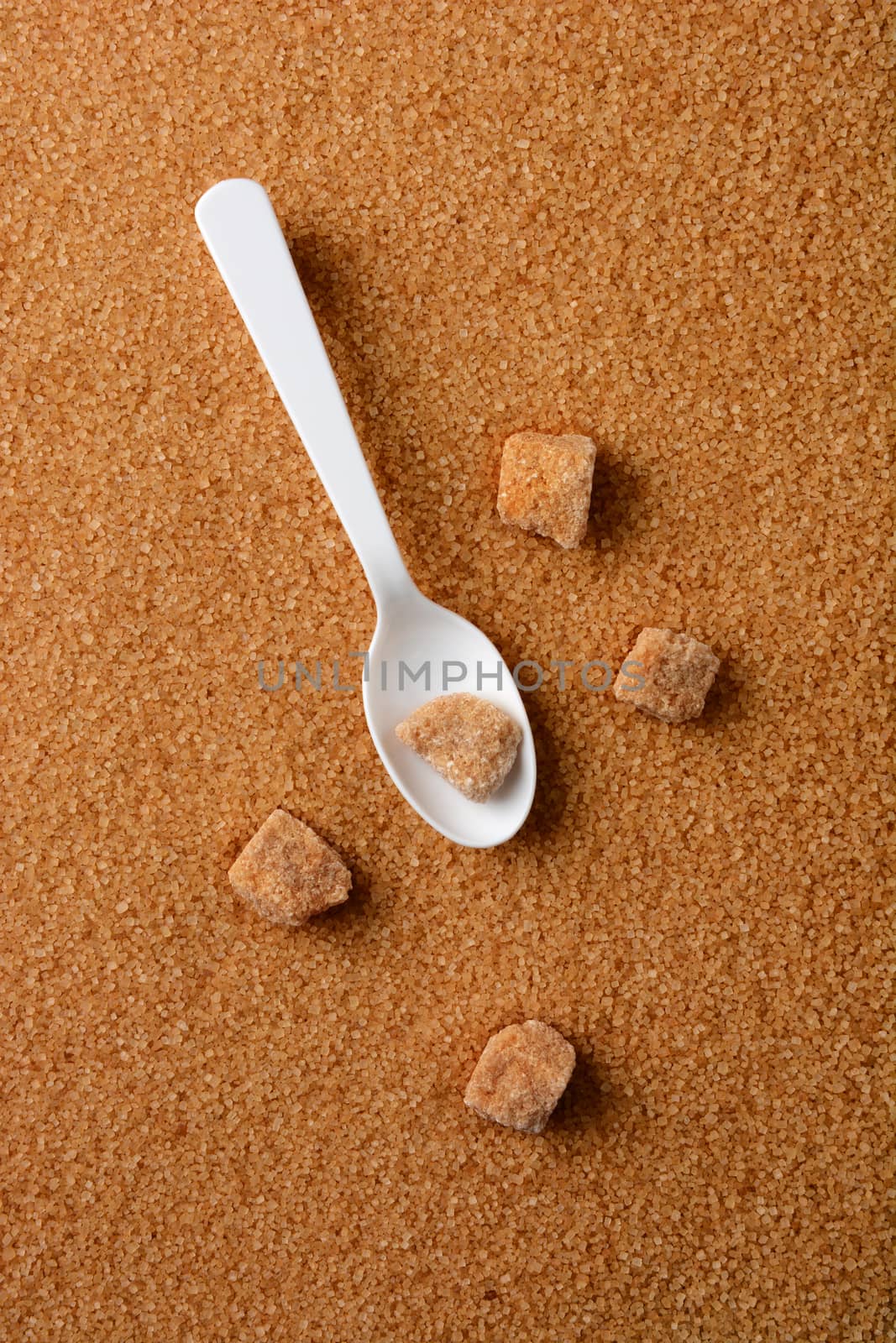 Overhead shot of lumps of raw brown sugar and a white spoon laying of a bed of granulated turinado sugar. Vertical format with copy space.